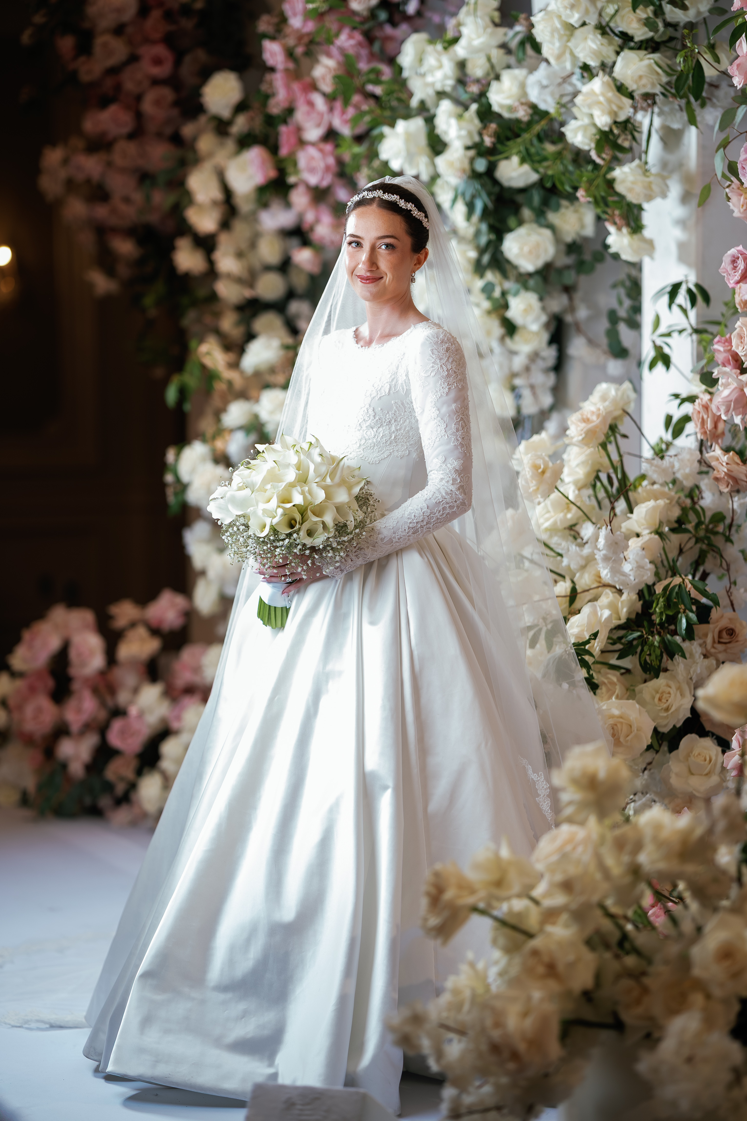 Bride in wedding dress and veil holding a bouquet at a floral ceremony backdrop with soft pastel flowers.