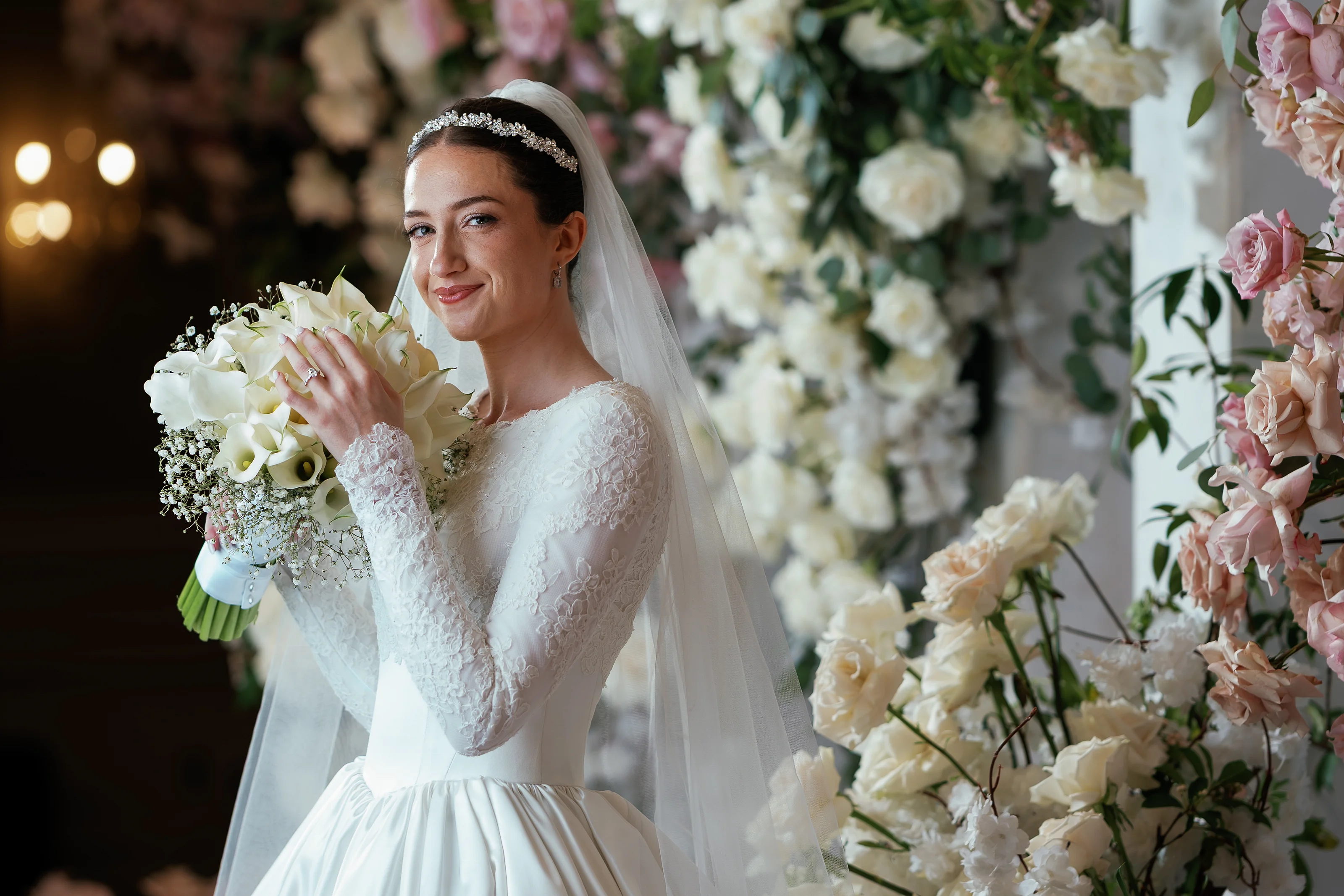 Bride in a white lace gown and veil smiling while holding a bouquet in front of an elegant floral wall.