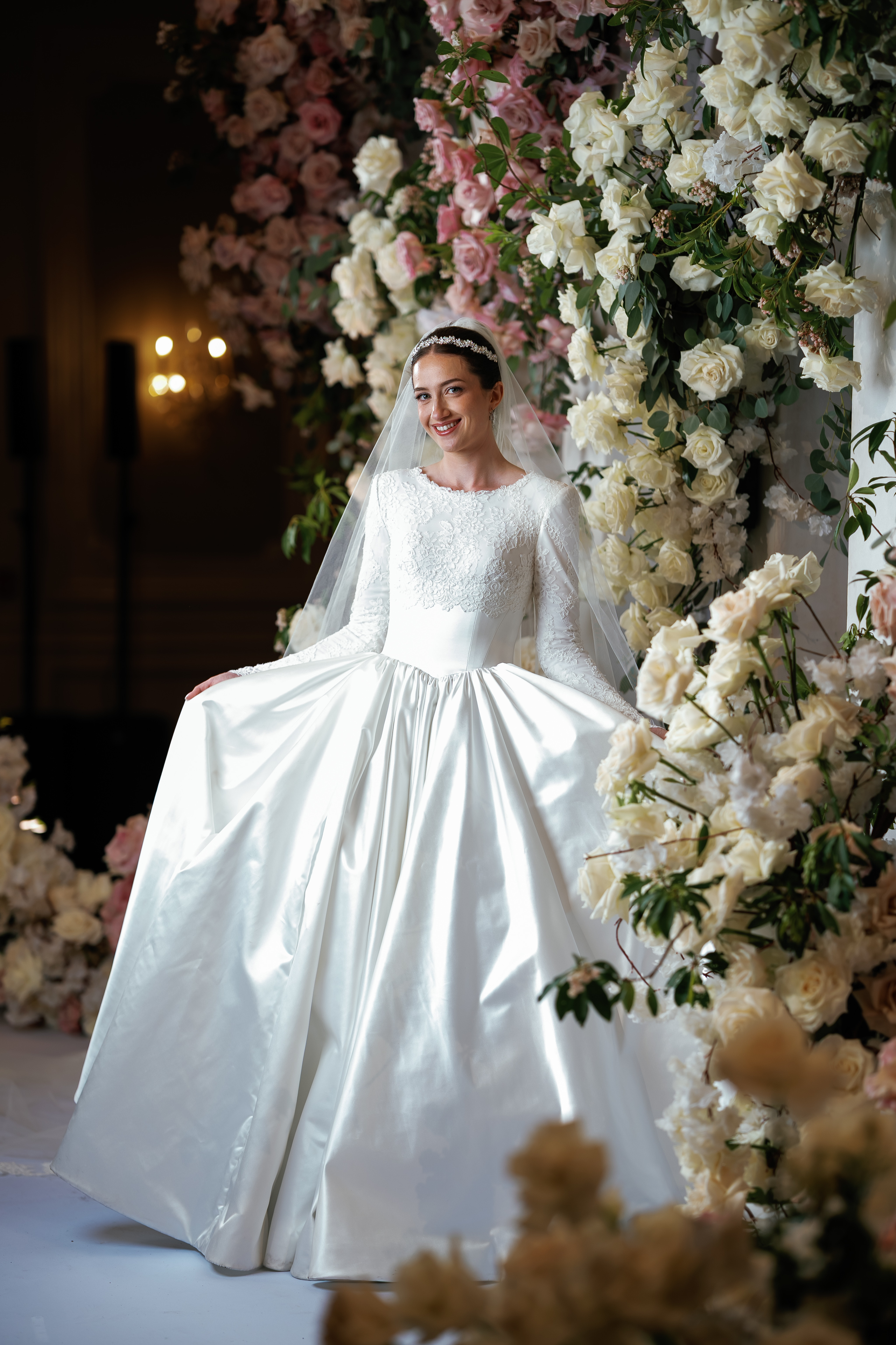 Bride in a white wedding gown and veil smiling in front of abundant cream and blush floral arrangements, warm indoor lighting and soft bokeh in the background.