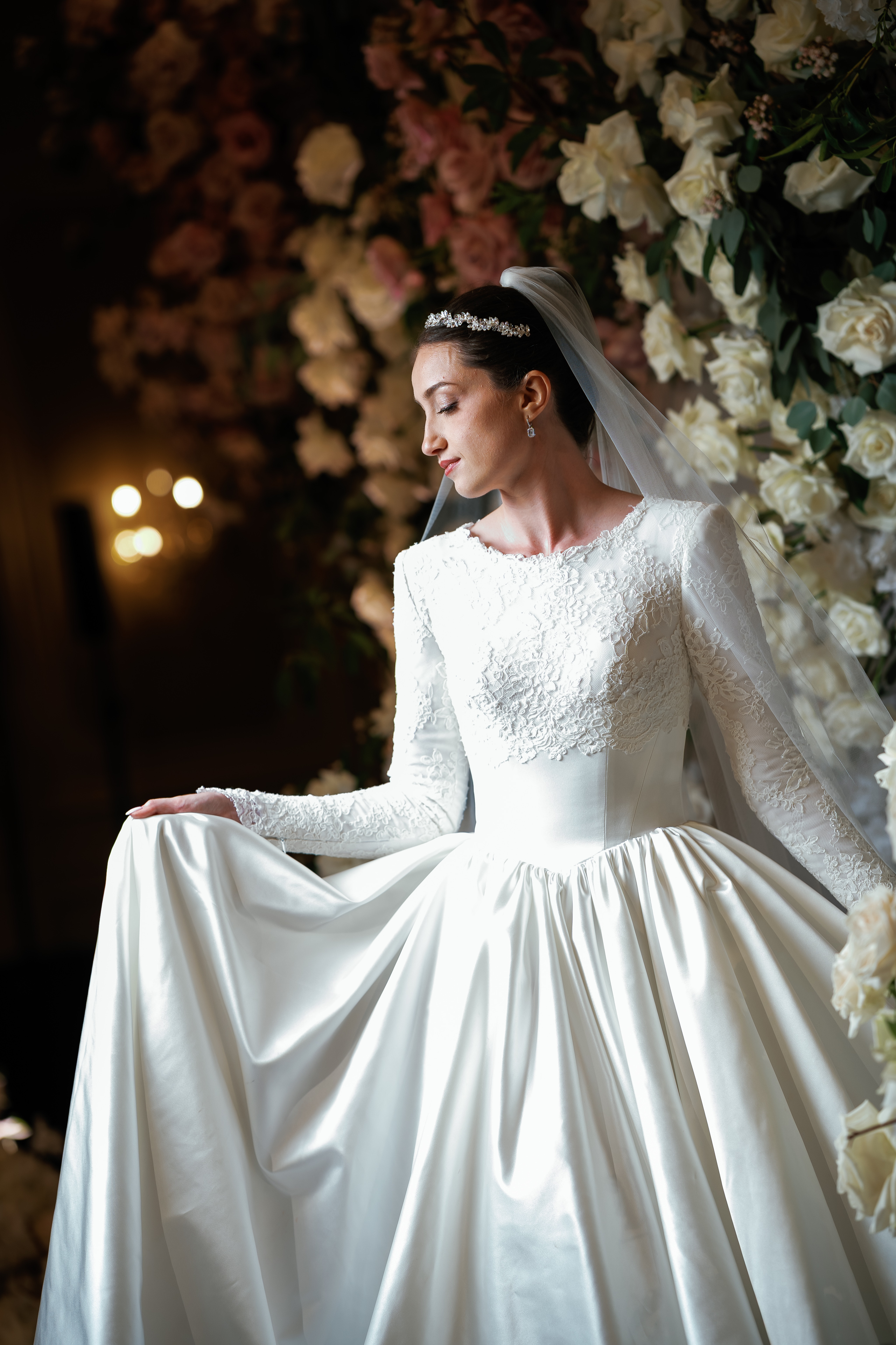 Bride in a white wedding gown and veil, eyes closed, posed beside a floral backdrop with warm bokeh lights in the background.