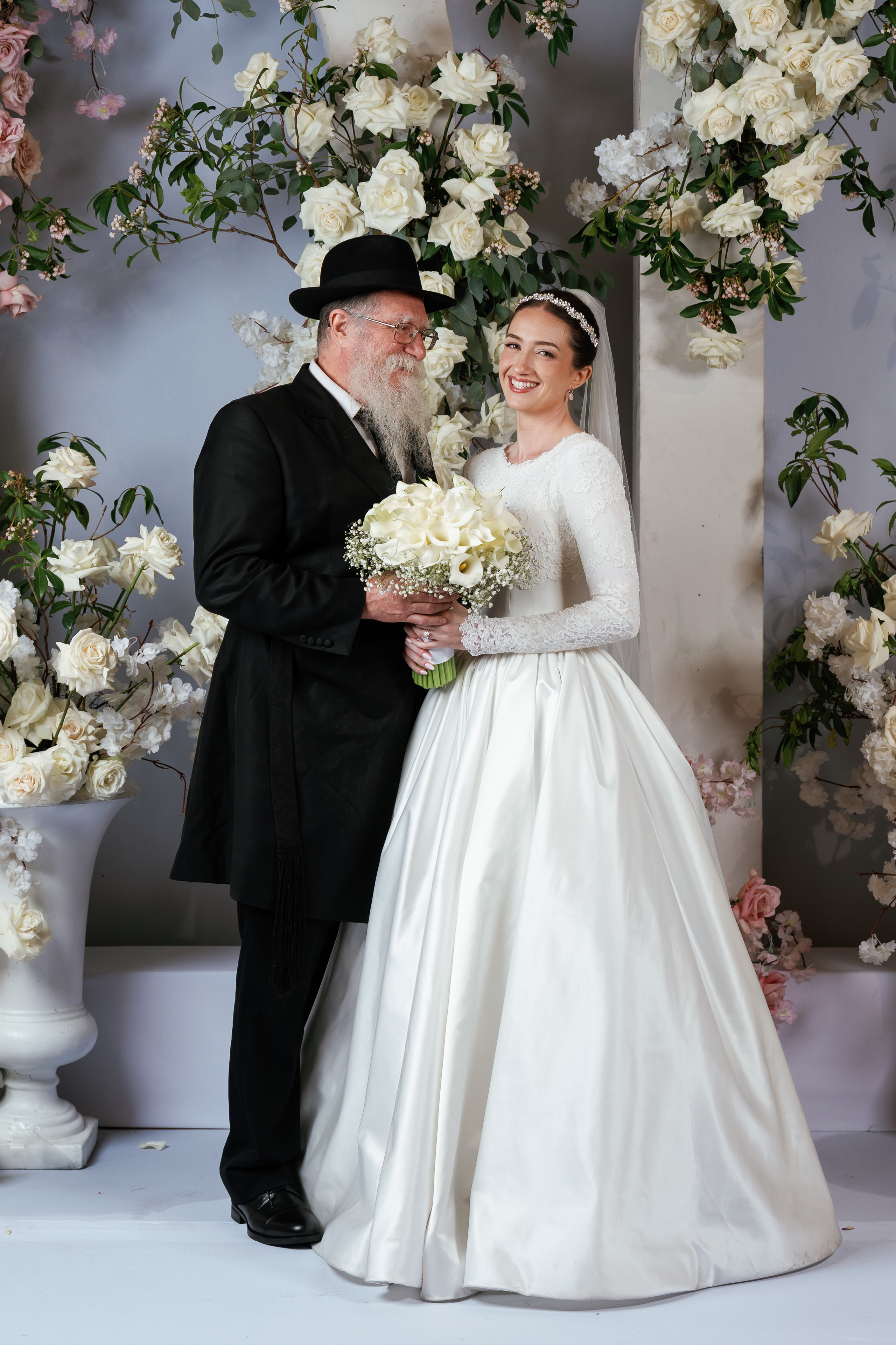 Smiling bride holding a bouquet with her groom during ceremony portraits against a floral backdrop of white roses and soft pink flowers.