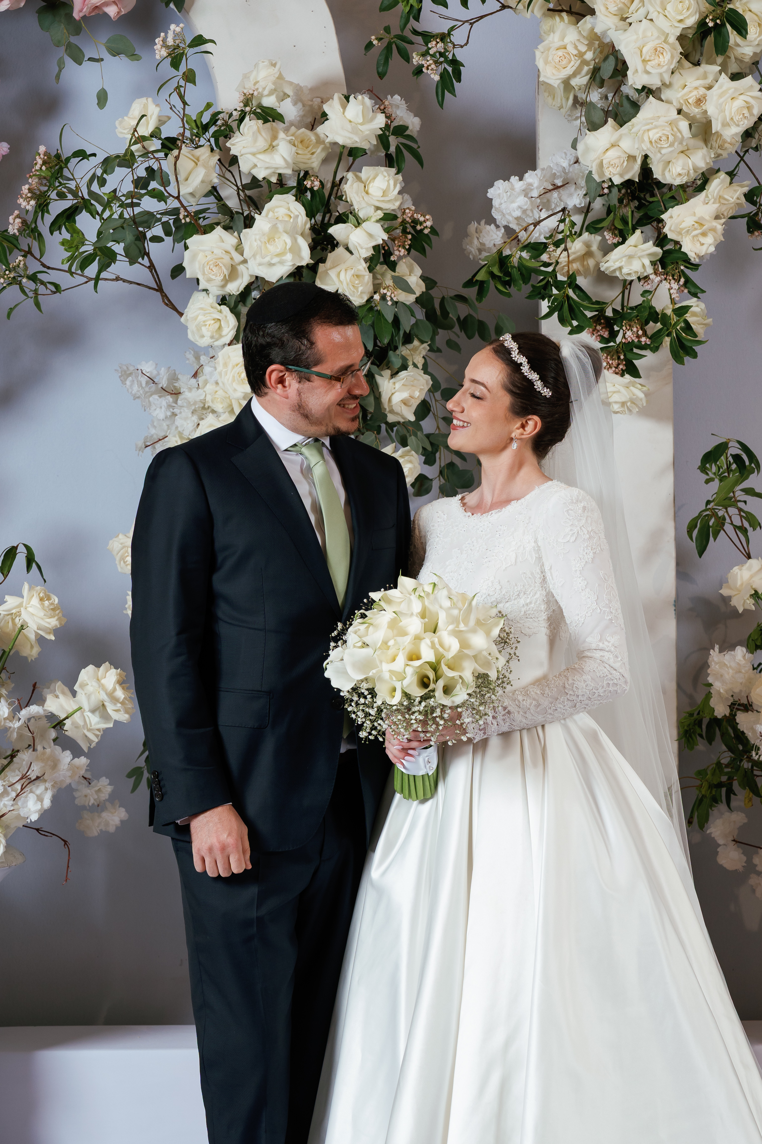 Smiling bride and groom standing under a white floral arch, bride holding a bouquet, gazing at each other.