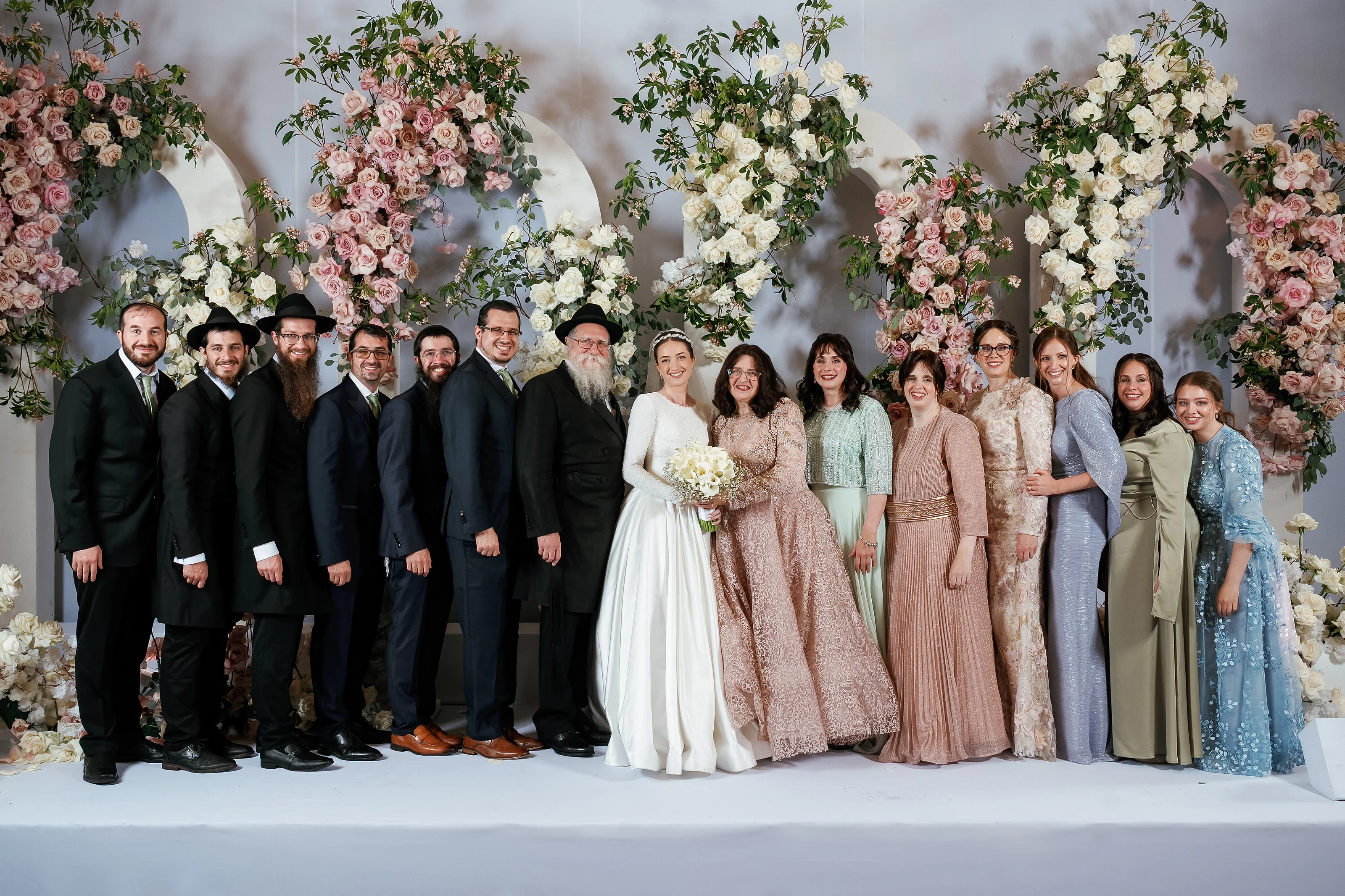 Wedding party and close family posed together on a bright stage, framed by pink and white floral arches.