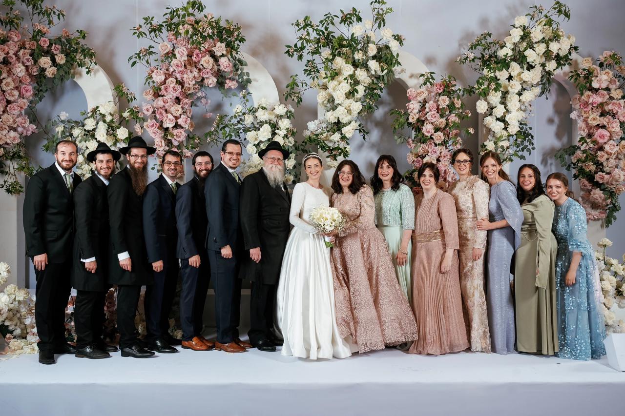 Wedding party and close family posed together on a bright stage, framed by pink and white floral arches. Wedding at Ateres Chynka, Brooklyn, NY.