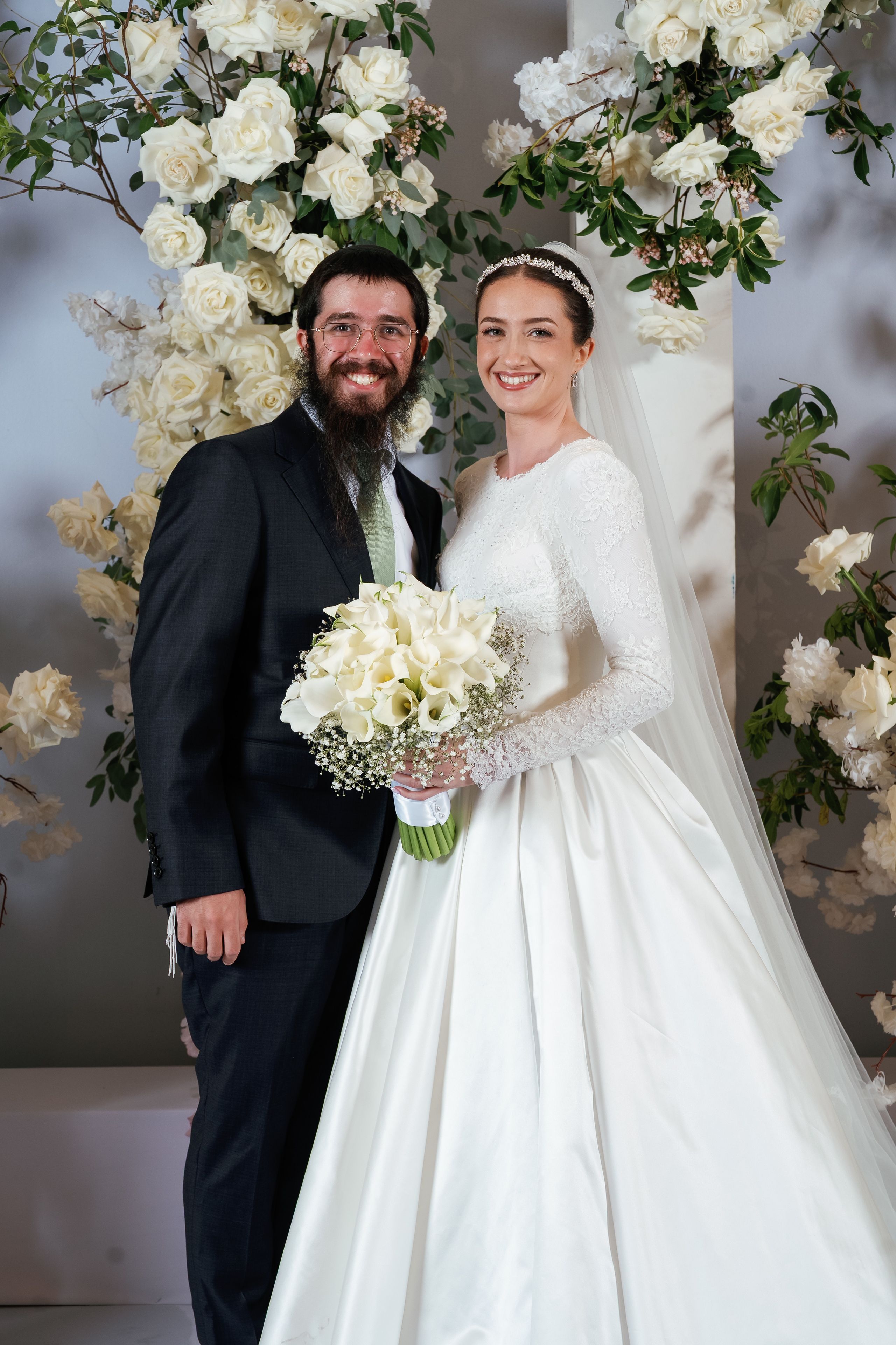 Bride and groom smiling together in front of an elegant floral backdrop, bride holding a bouquet with greenery accents.