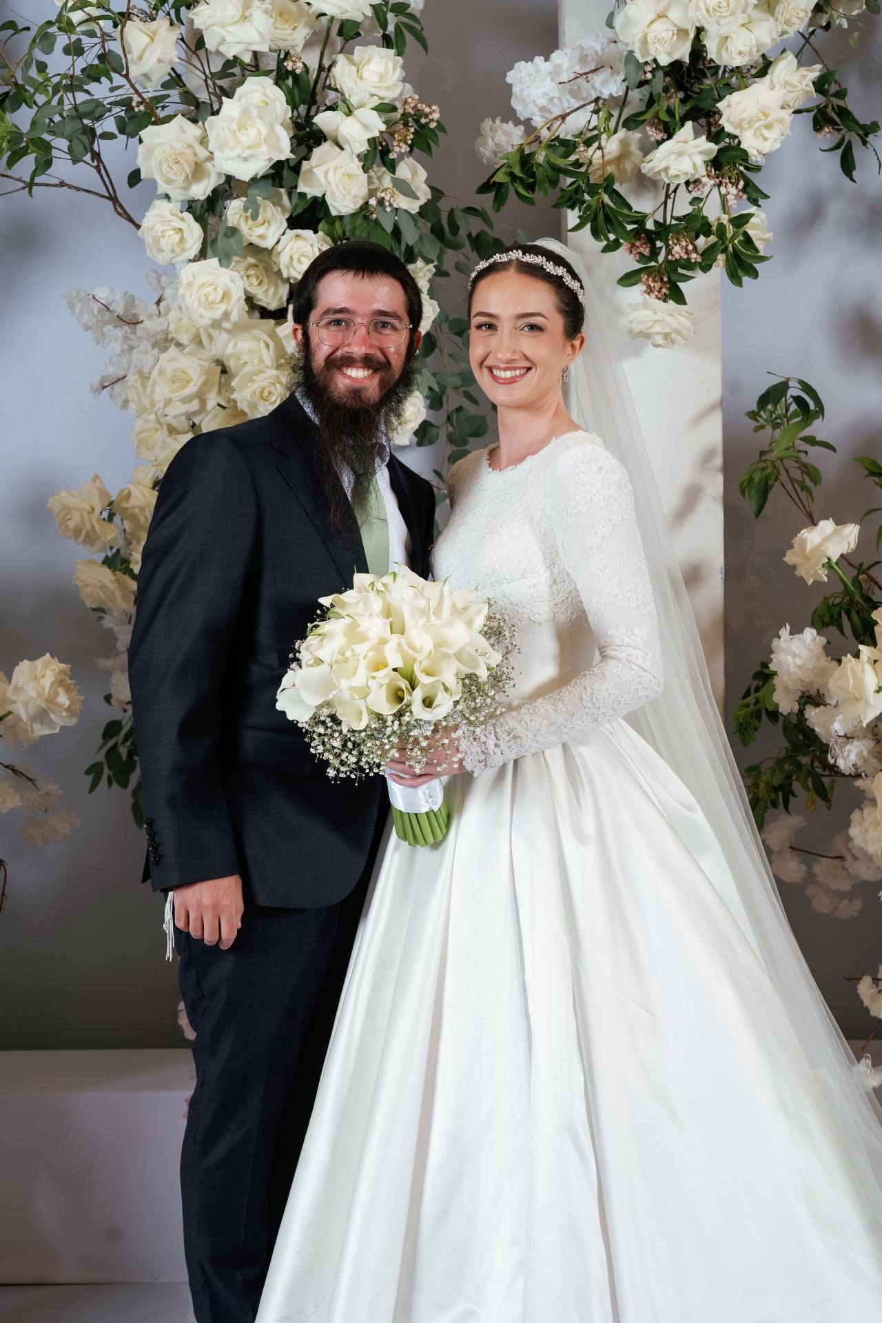 Bride and groom smiling together in front of an elegant floral backdrop, bride holding a bouquet with greenery accents. Wedding at Ateres Chynka, Brooklyn, NY.