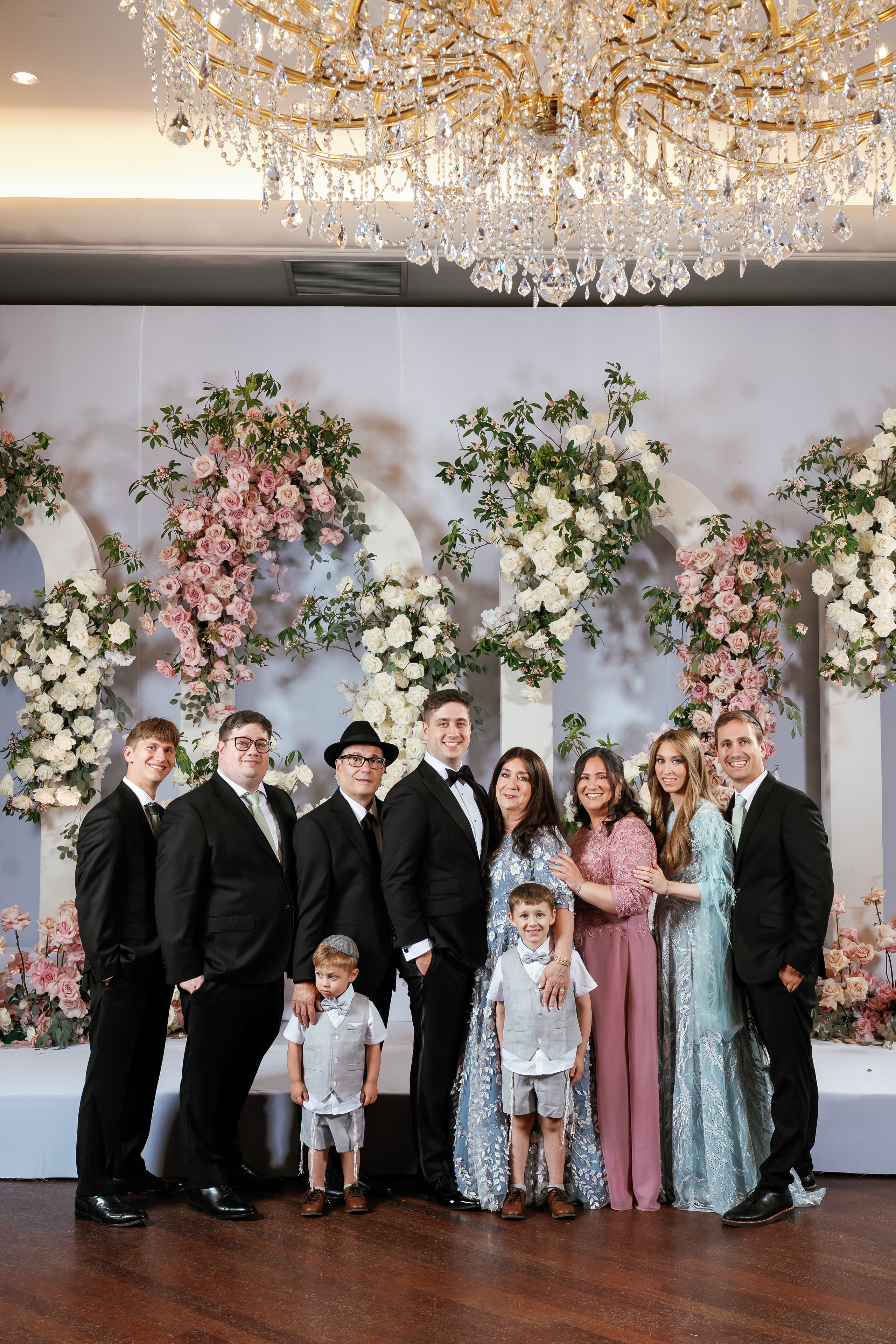 Posed group portrait of family and guests on a wedding stage with pink and white floral arches and a crystal chandelier overhead.