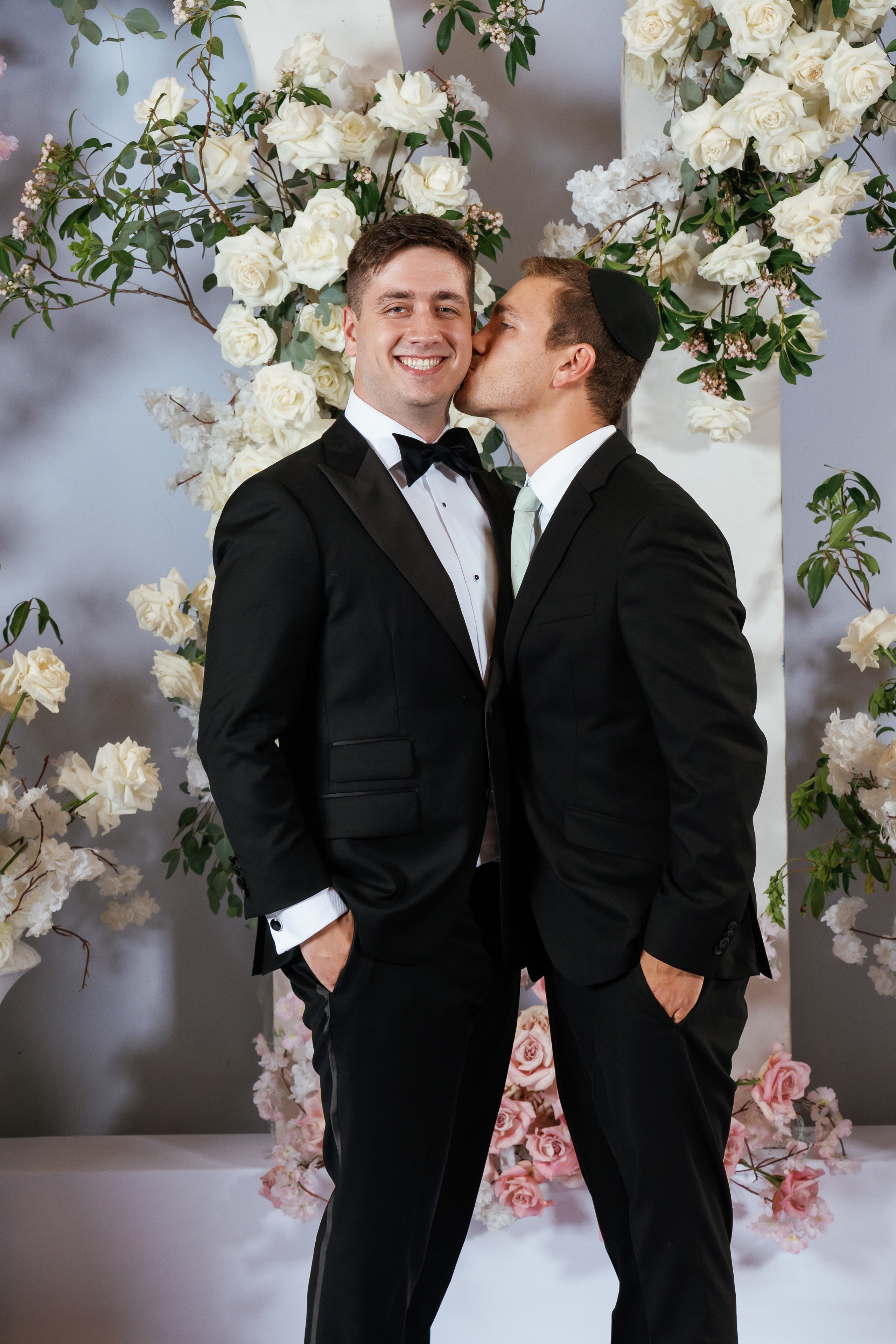 Smiling grooms sharing a kiss in front of a floral rose and greenery backdrop with elegant black formalwear