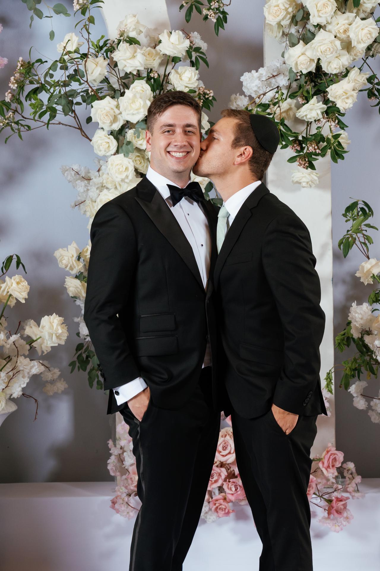 Smiling grooms sharing a kiss in front of a floral rose and greenery backdrop with elegant black formalwear. Wedding at Ateres Chynka, Brooklyn, NY.