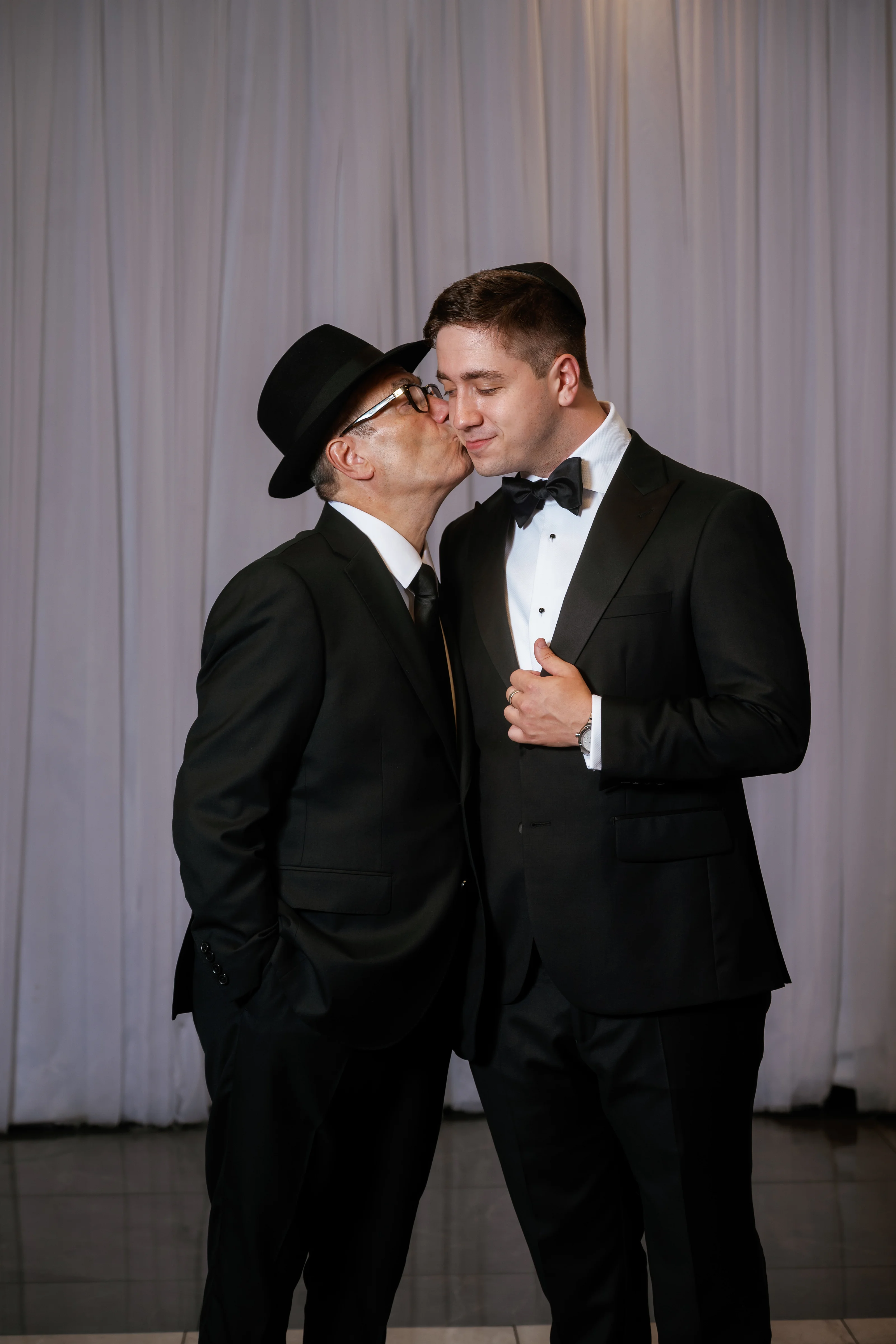 The groom leans in kissing his father during a private, intimate moment at a formal wedding celebration; both wear black tuxedos and bow ties in front of a draped backdrop.