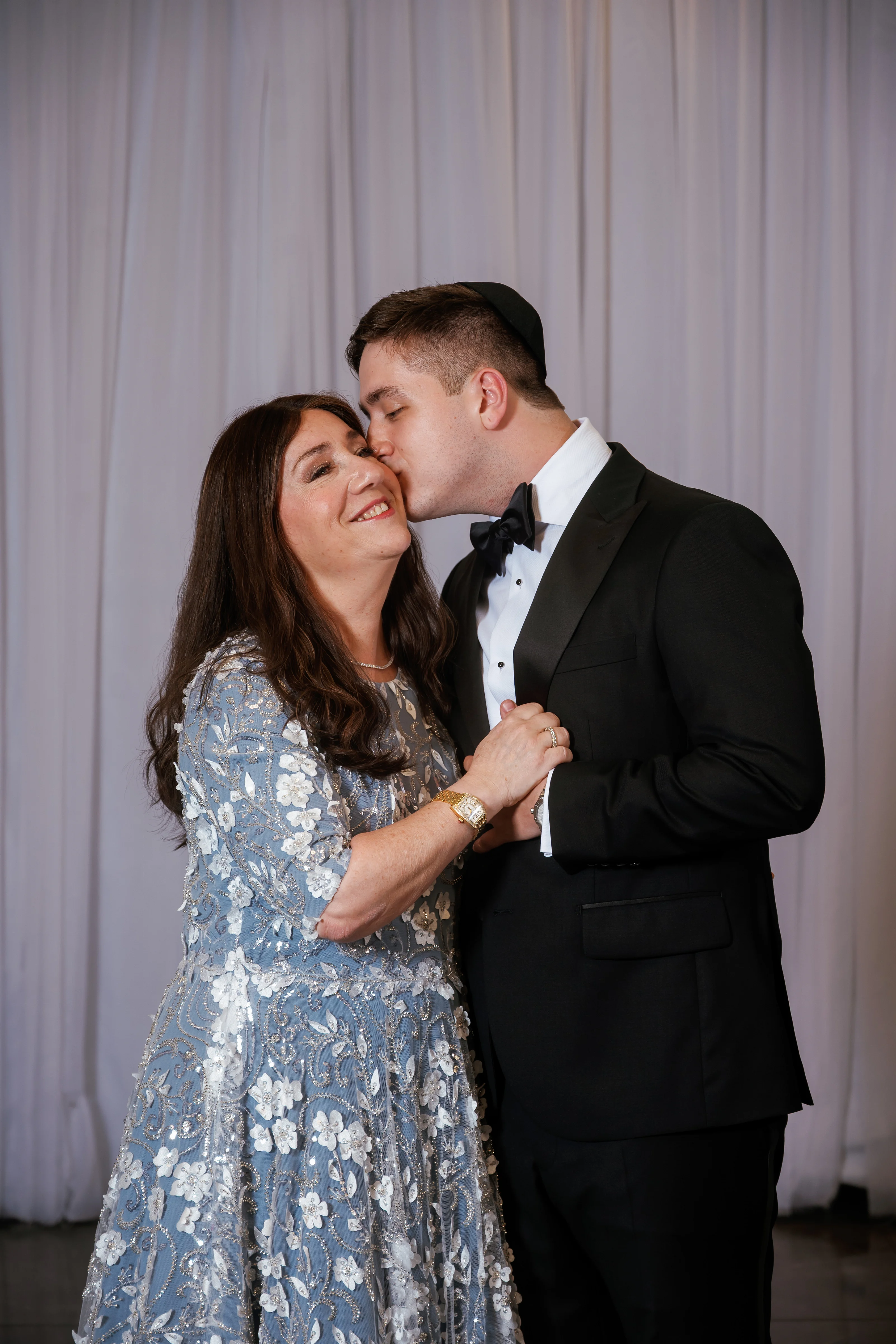 Bride and groom sharing a close embrace as the groom kisses the bride’s cheek in front of a draped backdrop.
