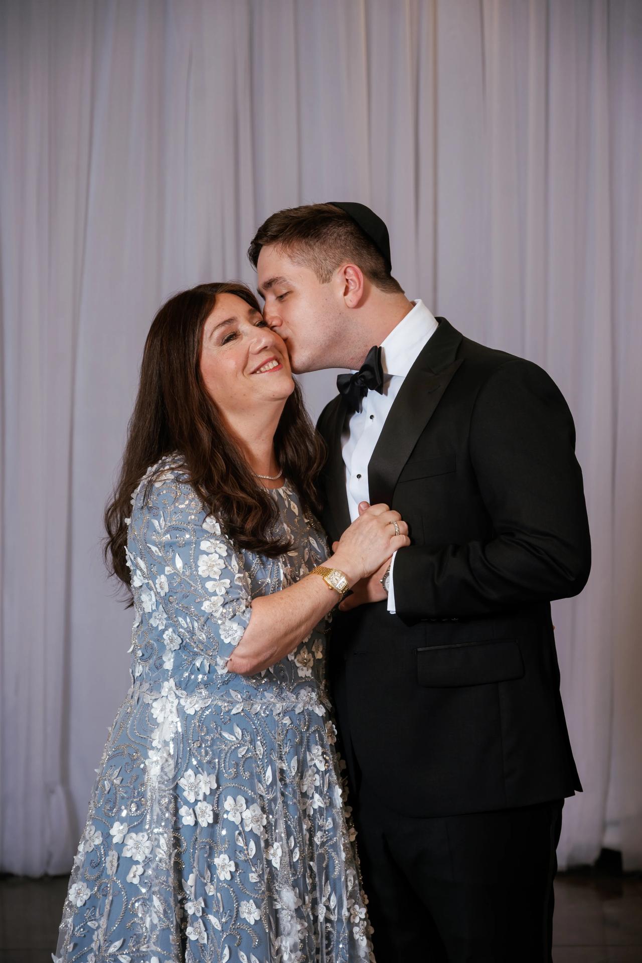 Bride and groom sharing a close embrace as the groom kisses the bride’s cheek in front of a draped backdrop. Wedding at Ateres Chynka, Brooklyn, NY.