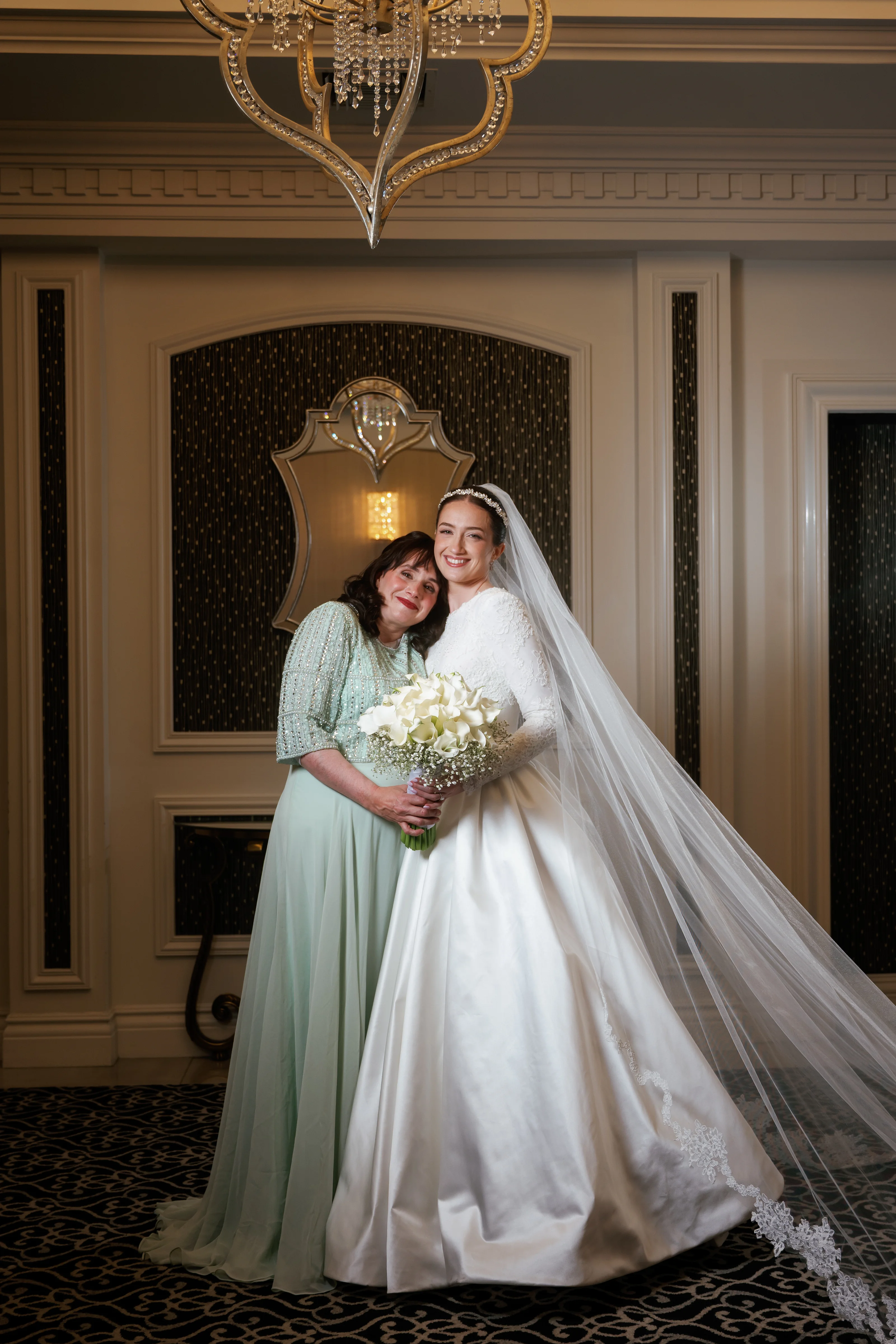 Bride in a white gown and veil hugging a loved one in an elegant ballroom setting with warm chandelier light.