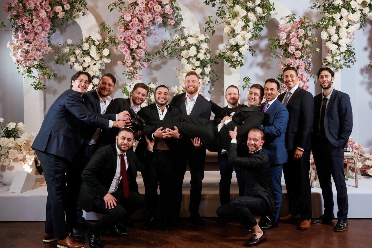 Posed group portrait of close family members in suits in front of large floral arches with pink and white roses. Wedding at Ateres Chynka, Brooklyn, NY.