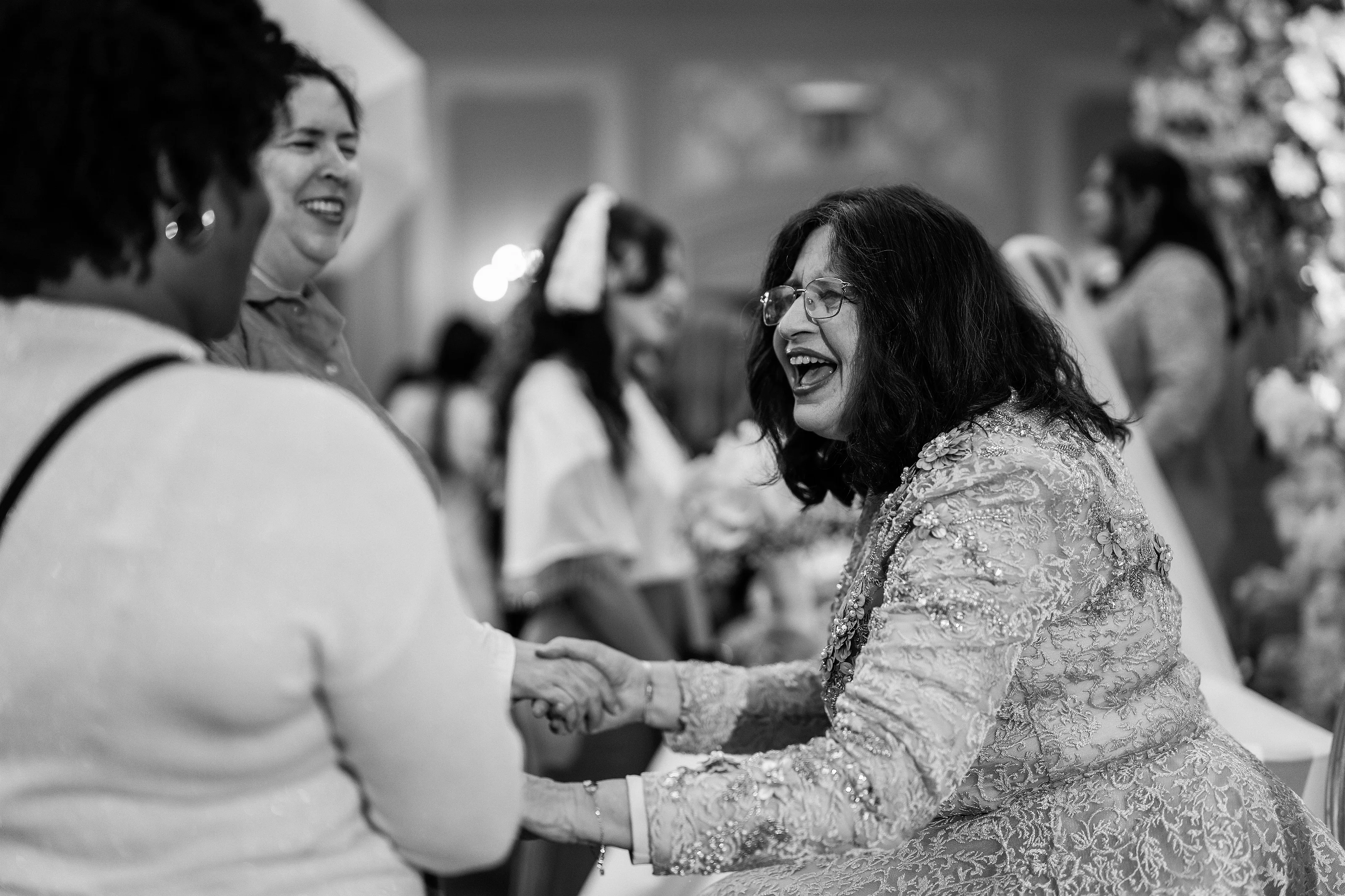 Black-and-white photo of a laughing woman in a lace dress during a joyous wedding celebration, sharing a warm moment with another guest.