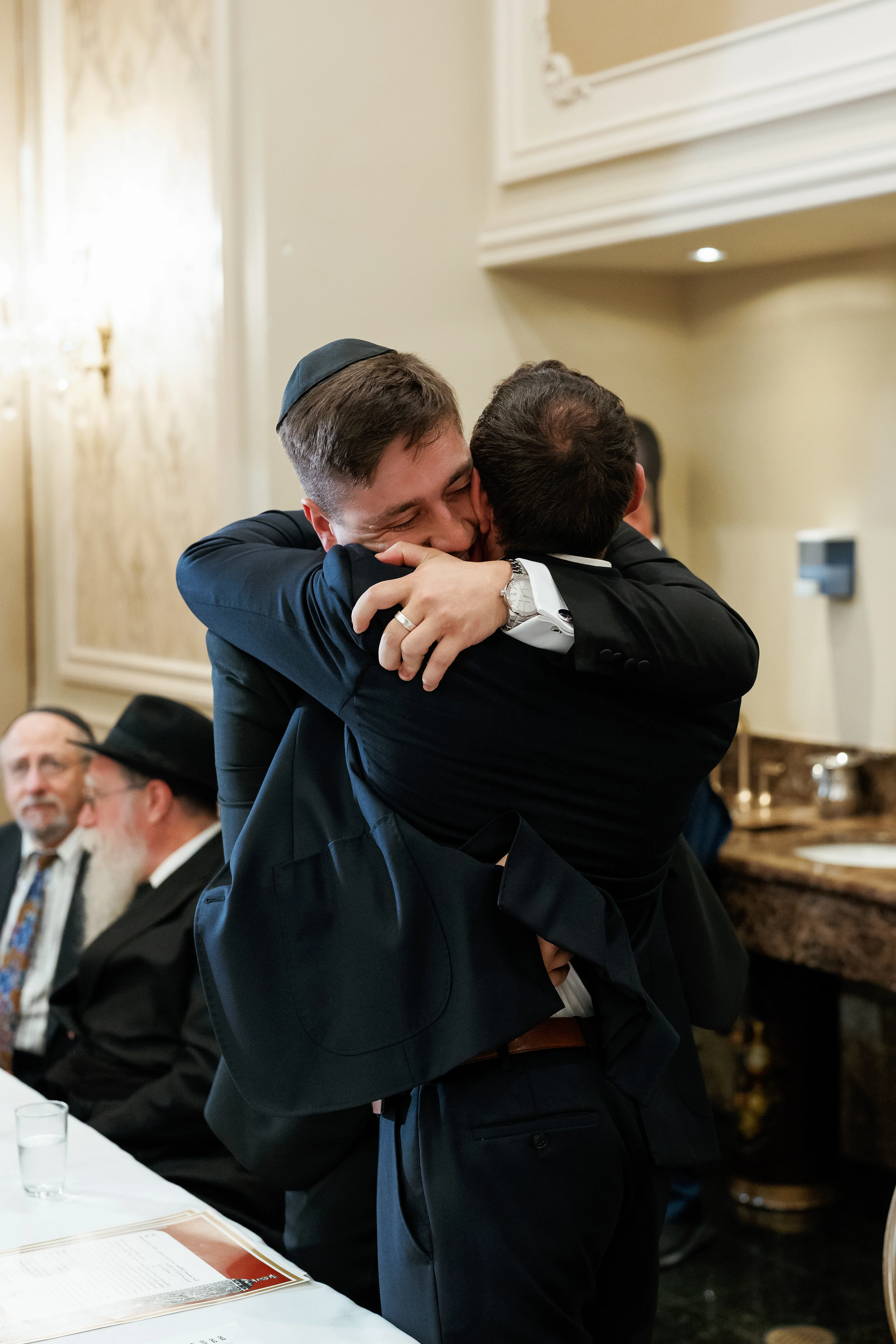 Two men in dark suits share a close embrace during a Jewish wedding ceremony moment, with other guests softly blurred in the background.
