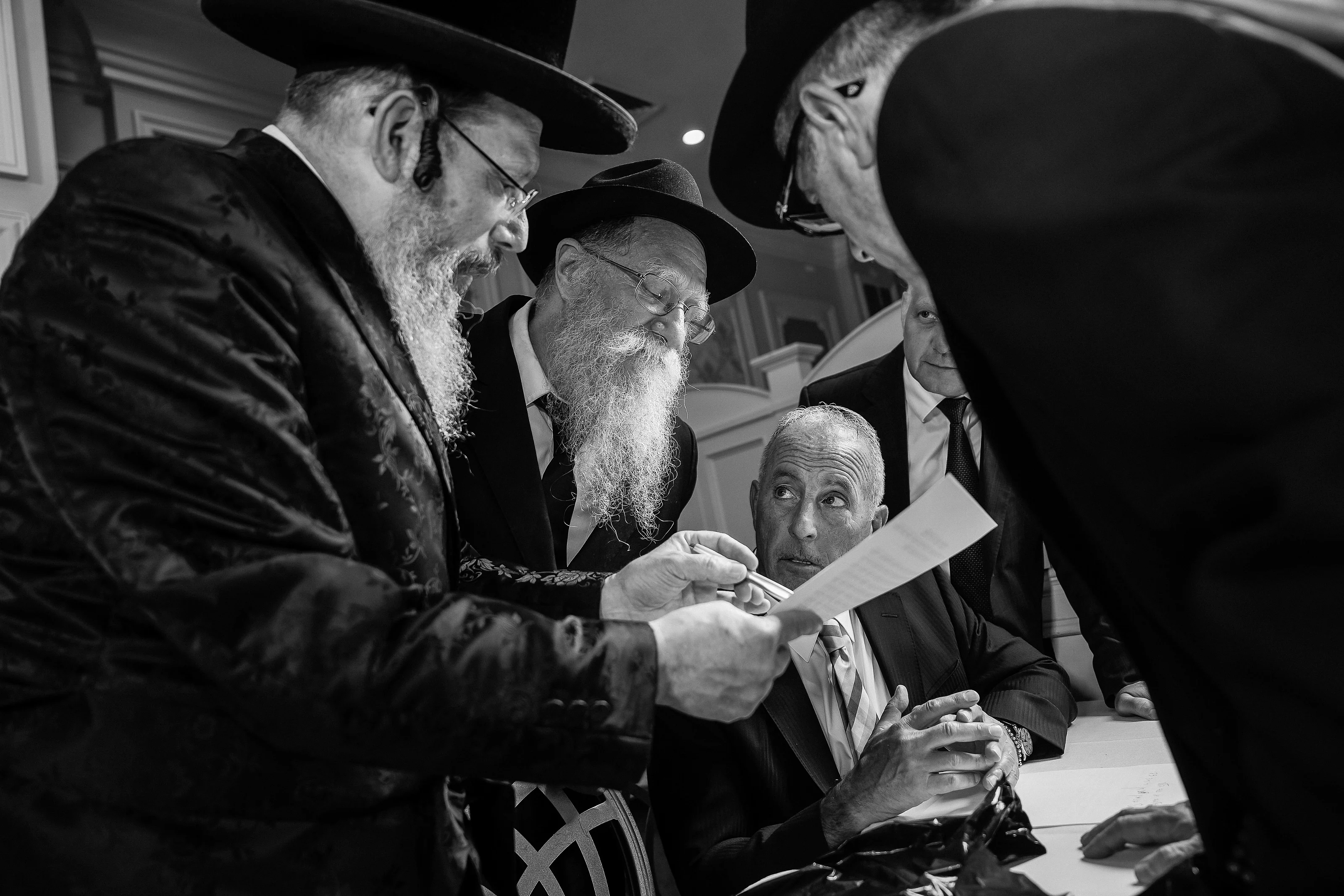 Black-and-white kabbalas-panim moment with men in hats leaning in, holding a small paper, and attentive faces gathered closely around the ritual.