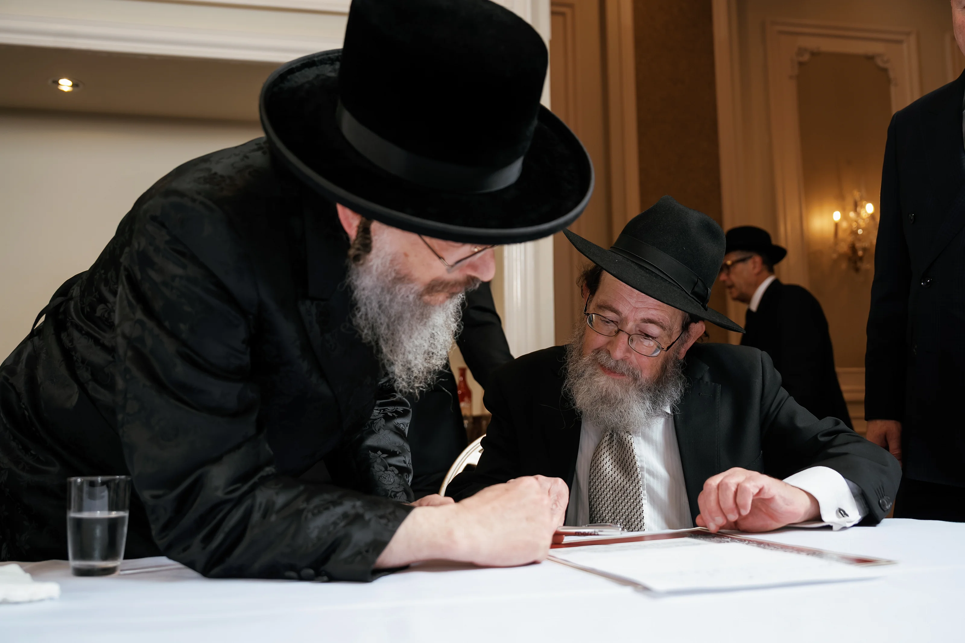 Two men in black hats lean in over documents on a table during a ceremony moment.