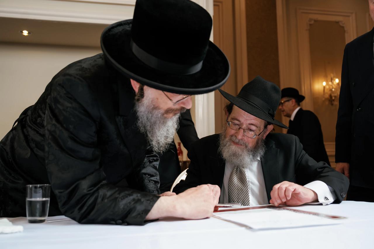 Two men in black hats lean in over documents on a table during a ceremony moment. Wedding at Ateres Chynka, Brooklyn, NY.