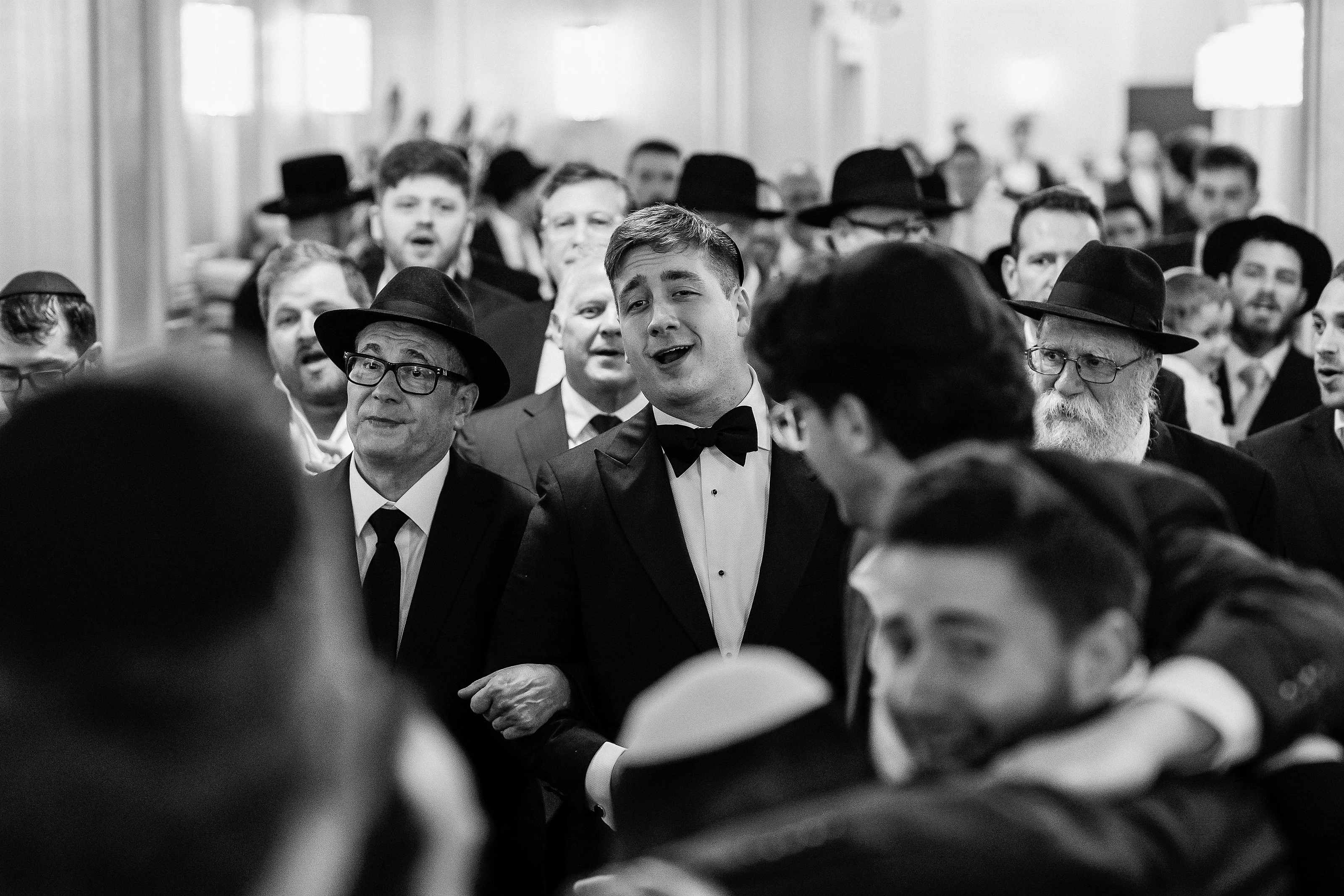 Black-and-white candid reaction moment during kabbalas-panim, groom centered with traditional guests in hats in the background.