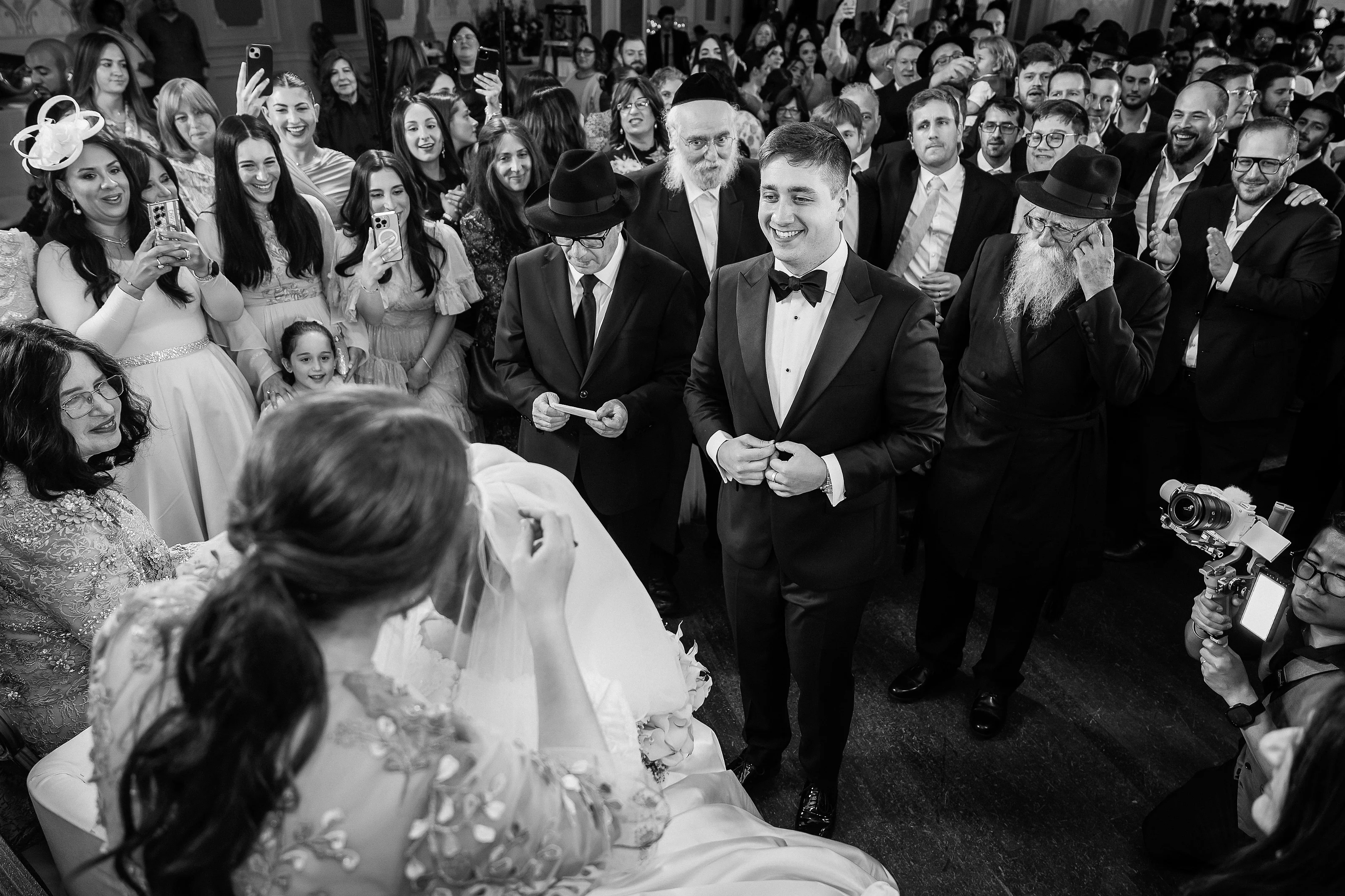 Black-and-white candid celebration moment with groom in tuxedo smiling while guests crowd together and phones raised; bride seen from behind in the foreground.