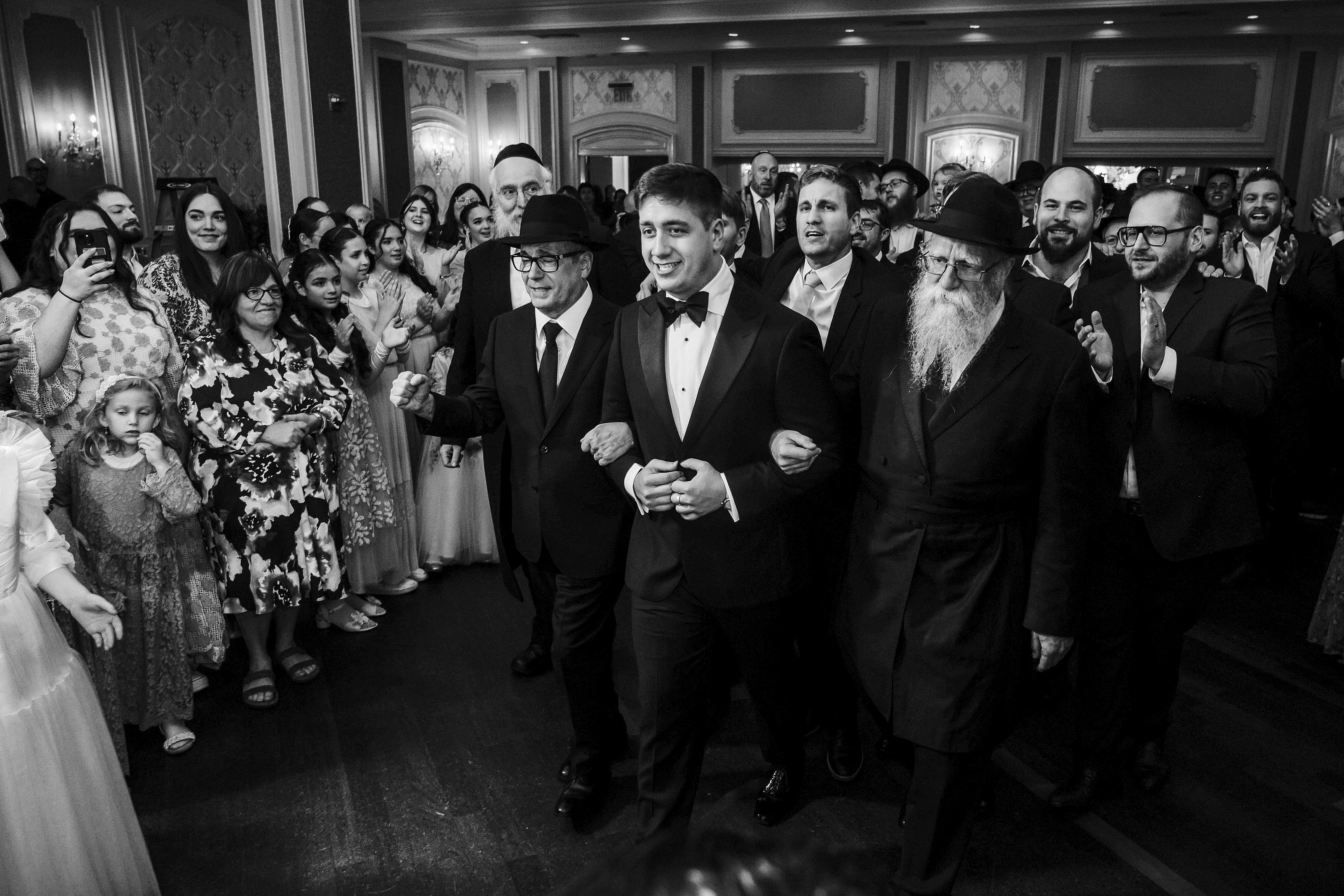 Black-and-white candid of a groom smiling while walking through a cheering reception crowd alongside a rabbi, with guests clapping in the background.