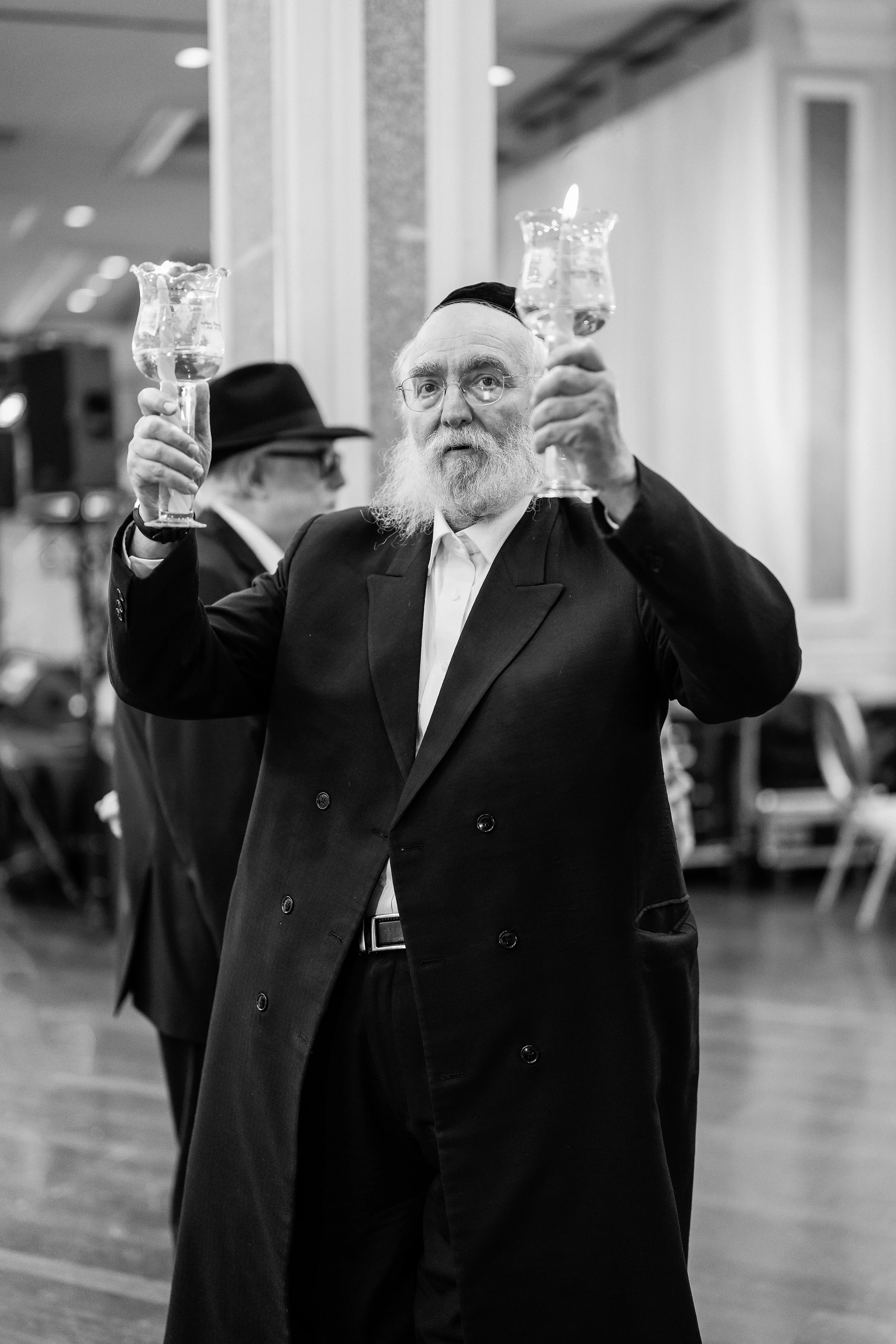 Black-and-white candid toast: a man in formal attire raises a champagne glass while confetti floats in a lit event hall, with another guest holding a glass behind him.