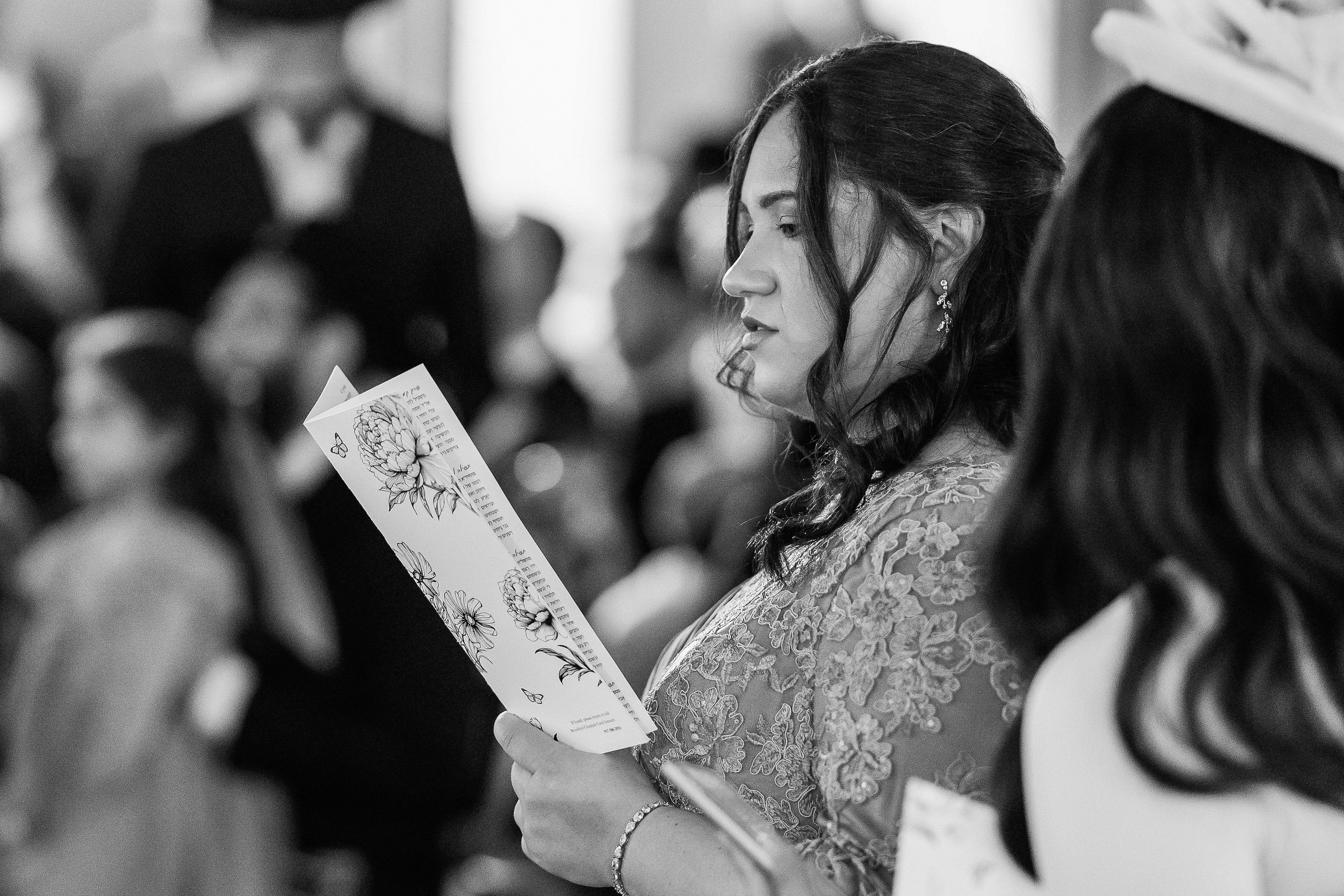 Black-and-white candid portrait of a bride in profile holding a printed ketubah reading card, surrounded by a softly blurred crowd.