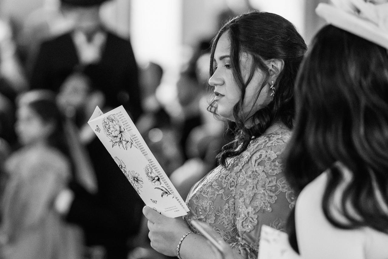 Black-and-white candid portrait of a bride in profile holding a printed ketubah reading card, surrounded by a softly blurred crowd. Wedding at Ateres Chynka, Brooklyn, NY.