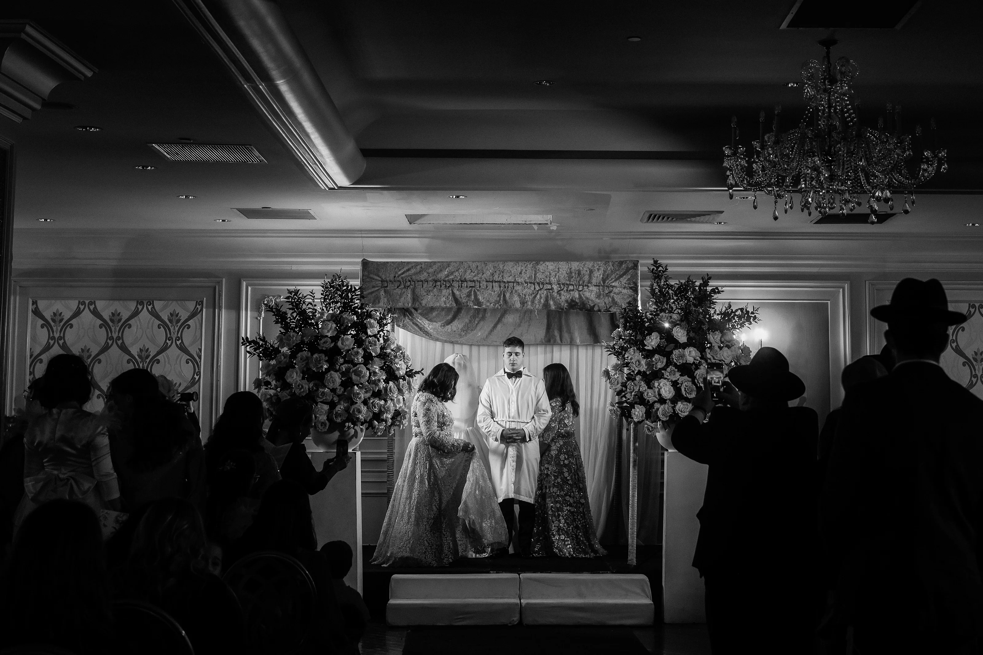 Black-and-white wide view of a wedding ceremony on a chuppah stage with floral arches and silhouetted guests in the foreground, chandelier overhead.
