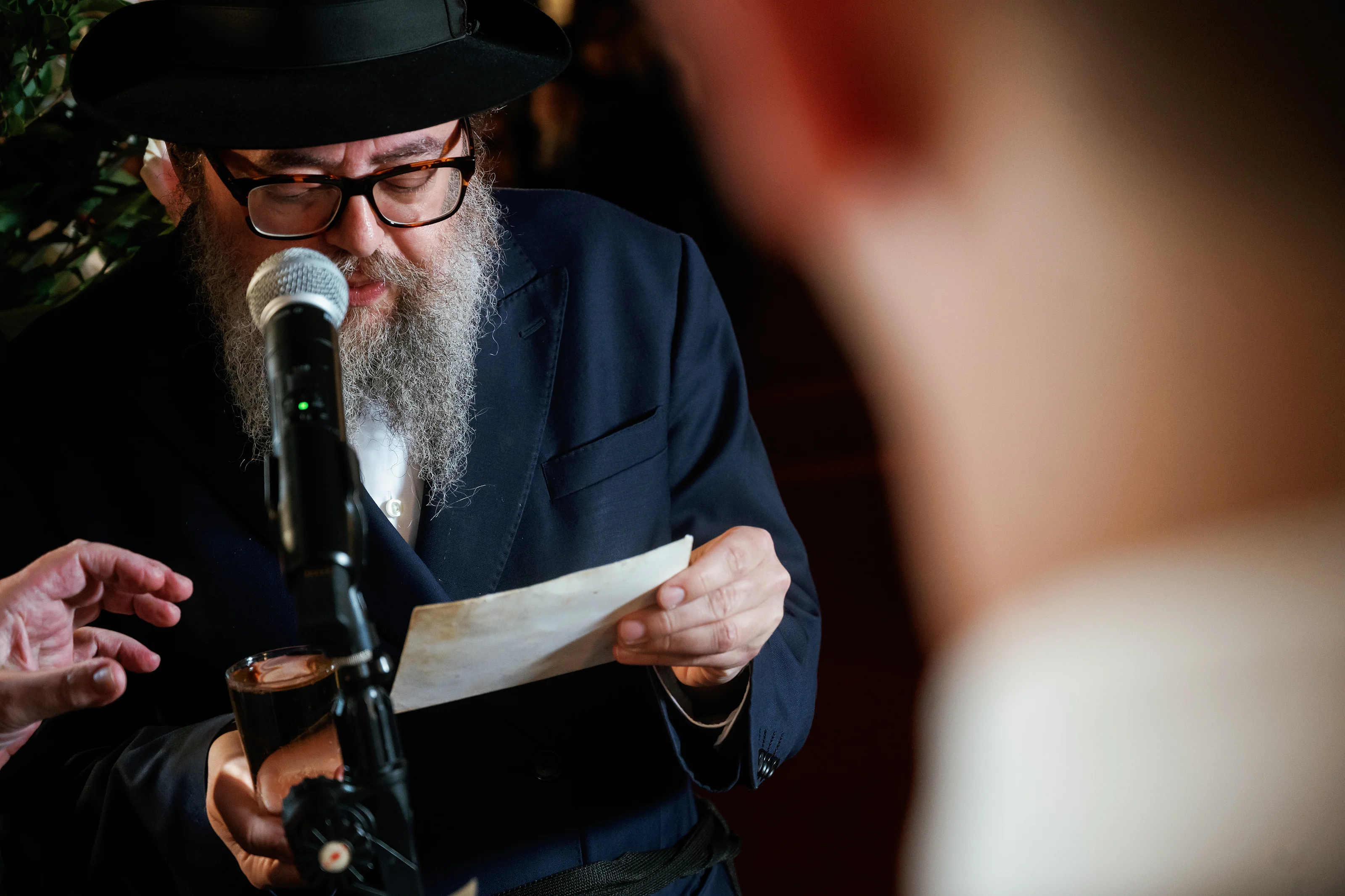 Man in a dark hat and suit reading from a note at a microphone during a formal Jewish event, with shallow depth of field and a blurred audience in the foreground.