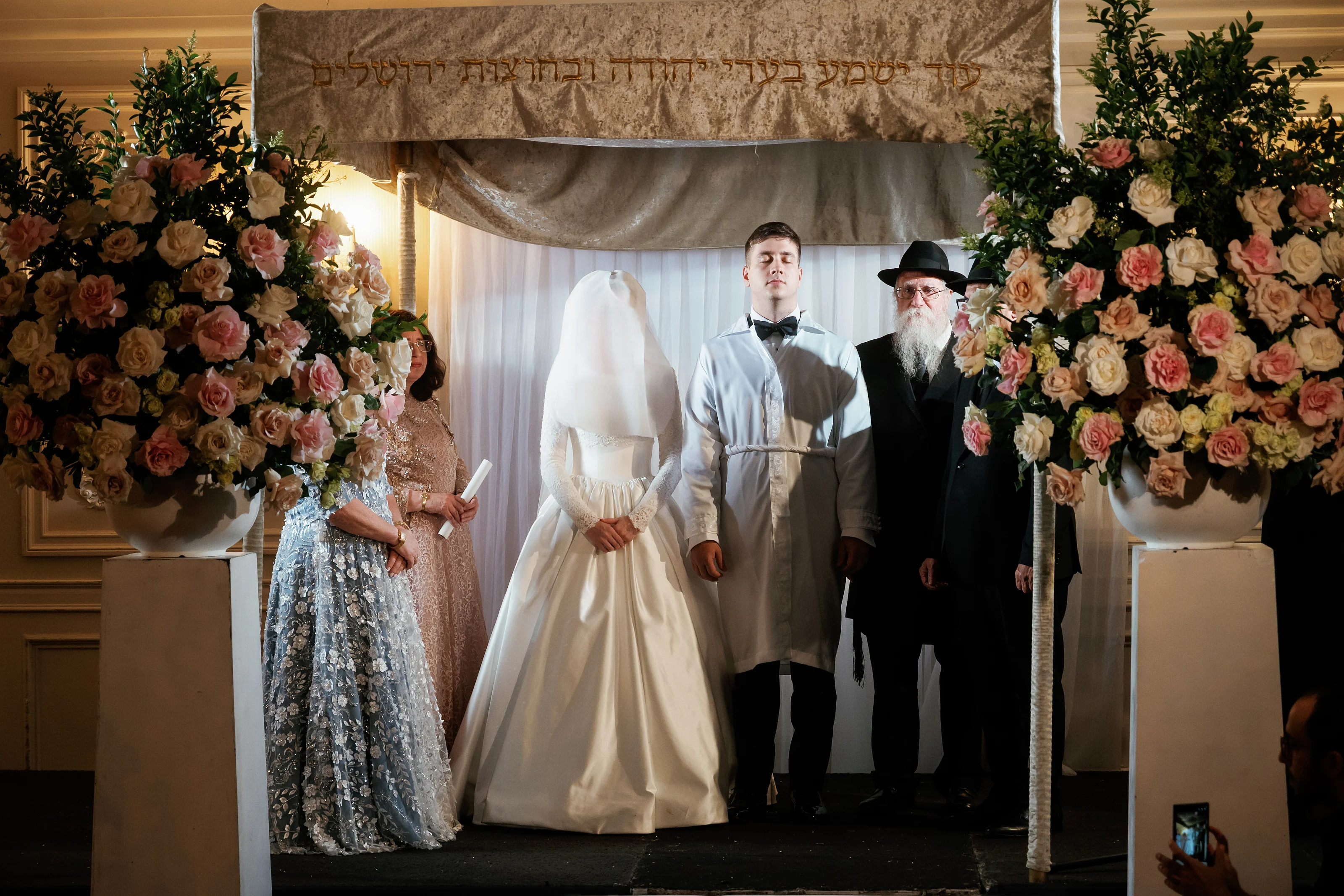 Bride in a white head covering and groom in a light suit standing under a at Ateres Chynka, photographed by CJ Studios
