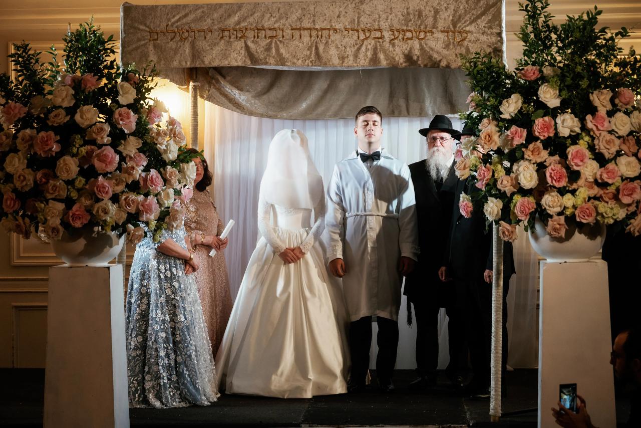 Bride in a white head covering and groom in a light suit standing under a at Ateres Chynka, photographed by CJ Studios