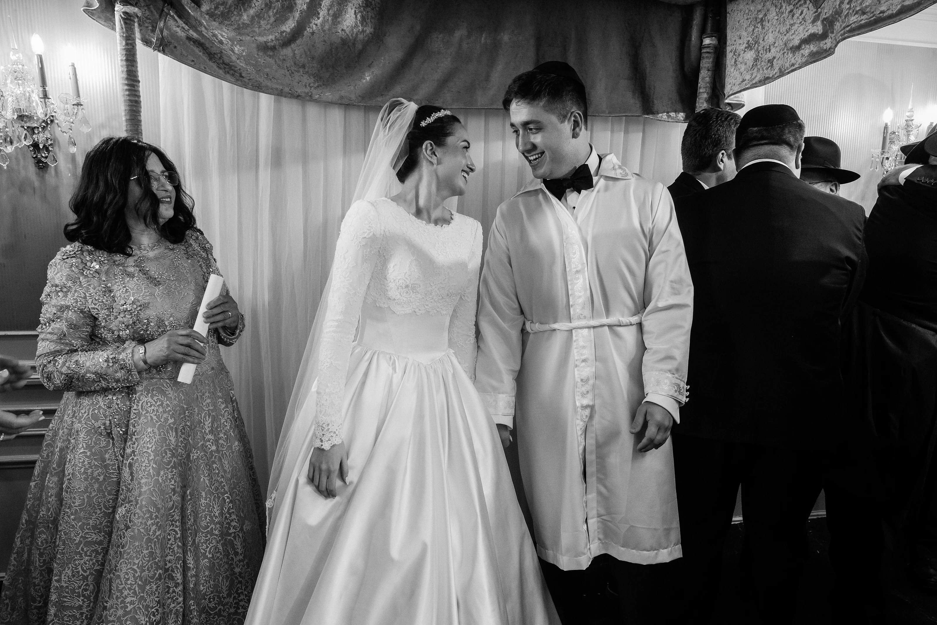Black and white wedding photo of bride and groom smiling during badeken as a veil drapes over the bride’s face, with family watching nearby.