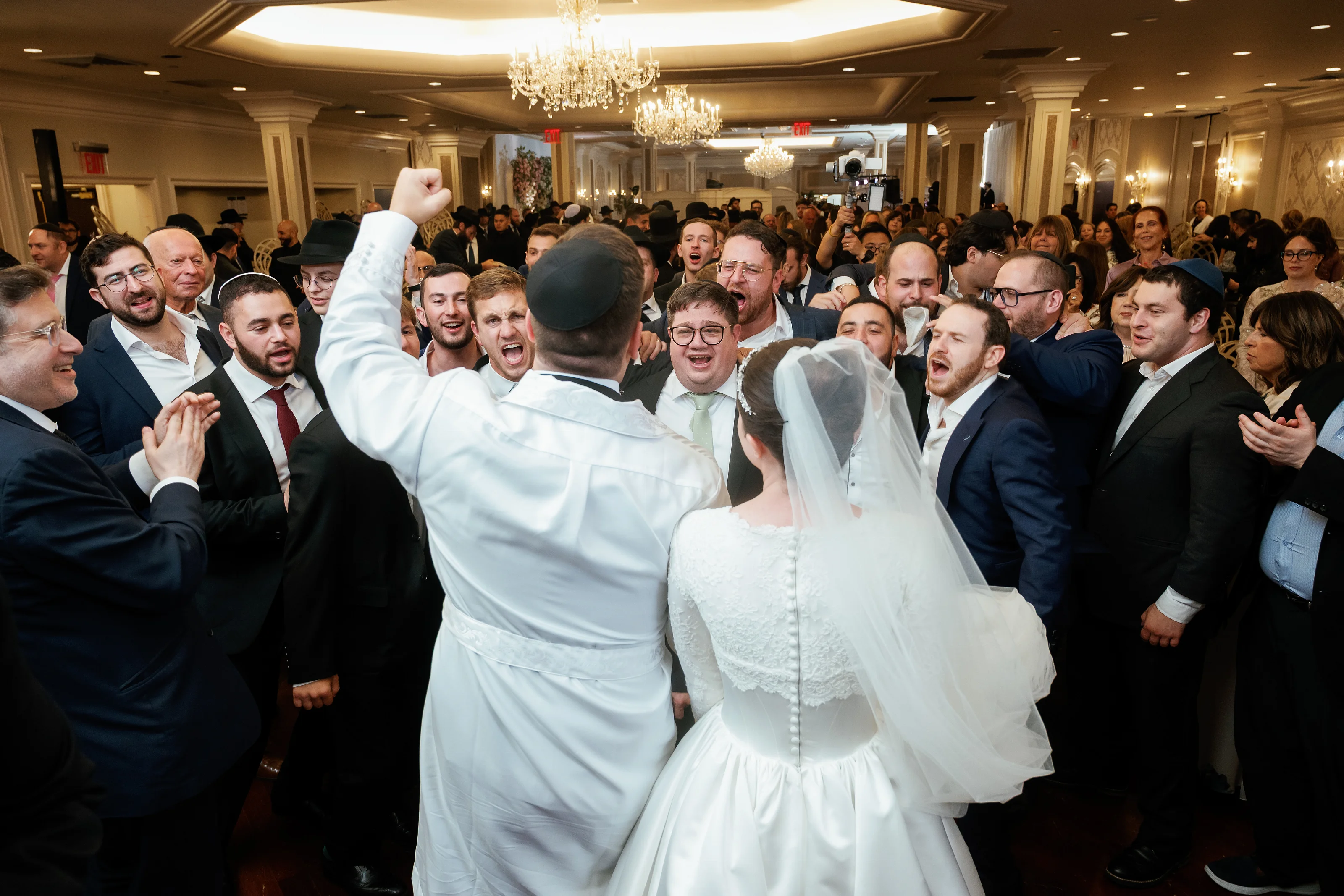 Groom and bride in the center of a crowded ballroom during a joyous reception moment, surrounded by friends cheering and celebrating.