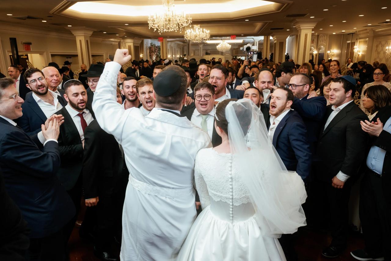 Groom and bride in the center of a crowded ballroom during a joyous reception moment, surrounded by friends cheering and celebrating.