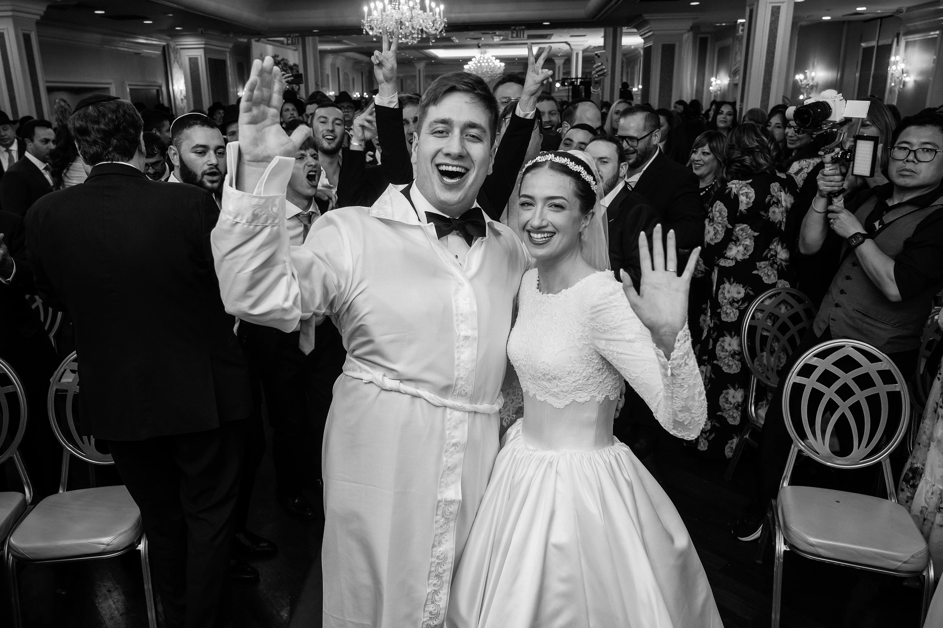Black-and-white candid of a wedding couple dancing and laughing with hands raised at an indoor reception, crowd watching and cheering behind them.