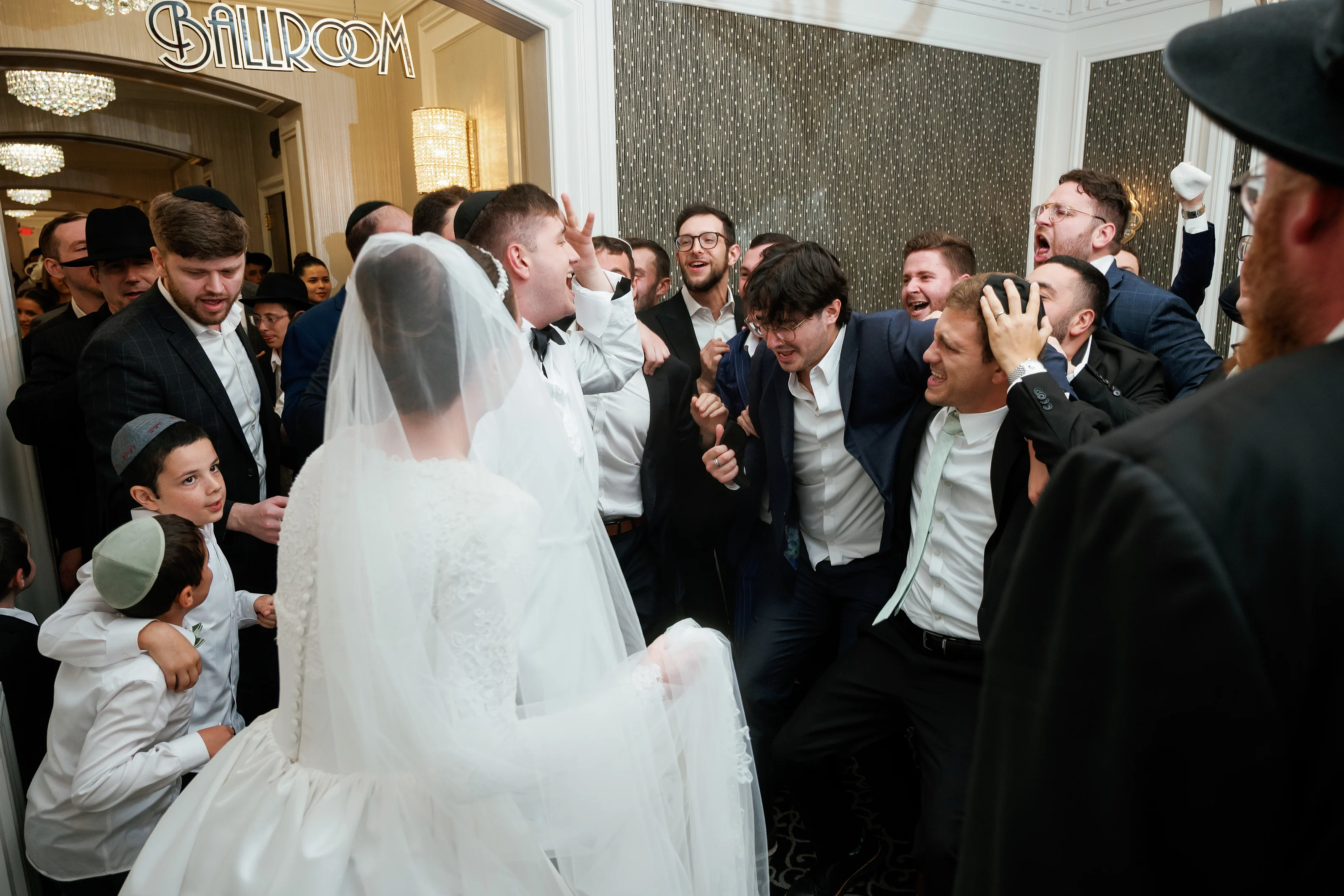 Bride in a white veil turns toward a cheering group in a chandelier-lit reception hall as guests laugh and react during the celebration.