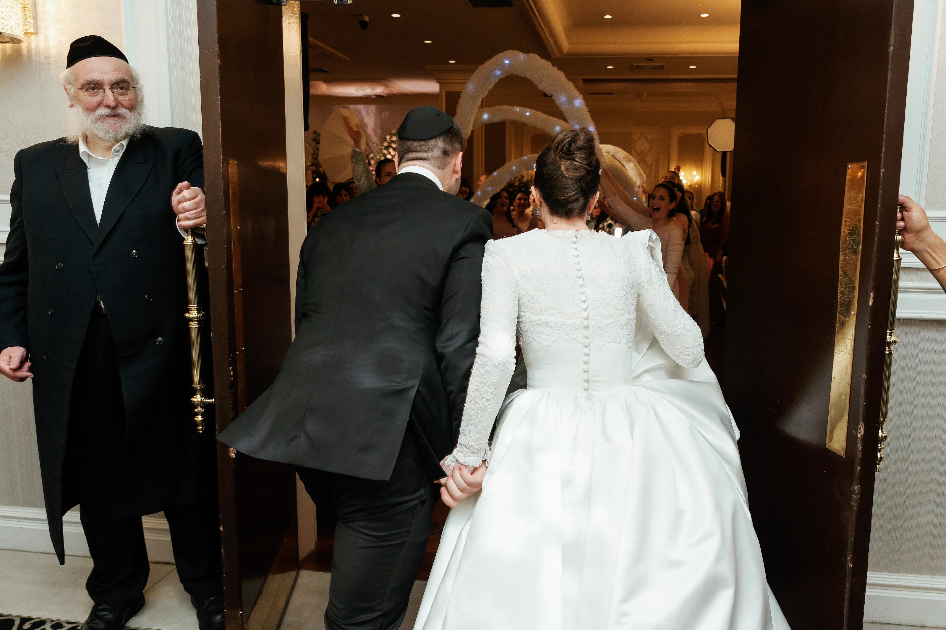 Bride and groom holding hands in a doorway moment during a Jewish wedding reception, warm indoor lighting and guests in the background.