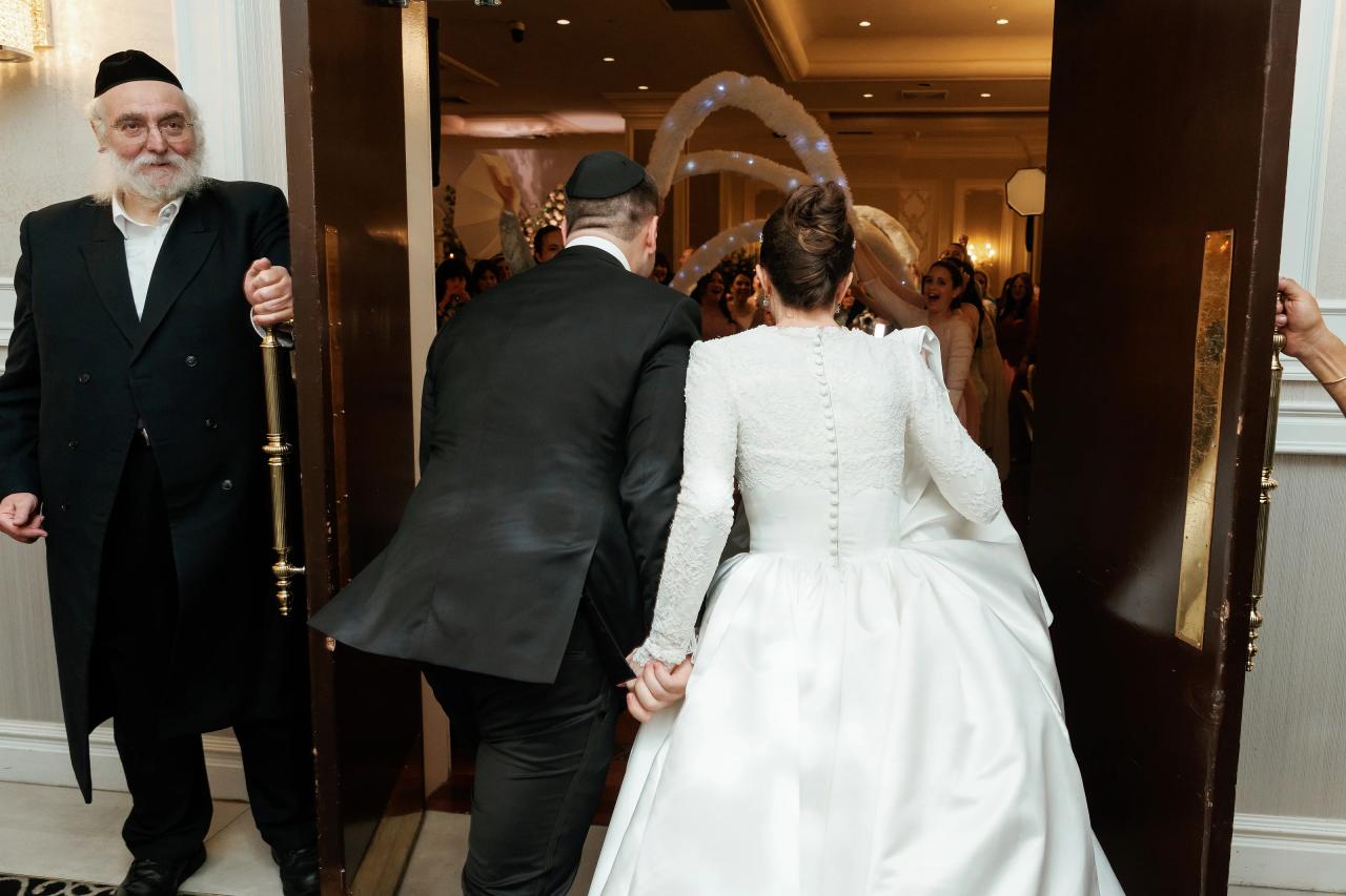 Bride and groom holding hands in a doorway moment during a Jewish wedding reception, warm indoor lighting and guests in the background. Wedding at Ateres Chynka, Brooklyn, NY.