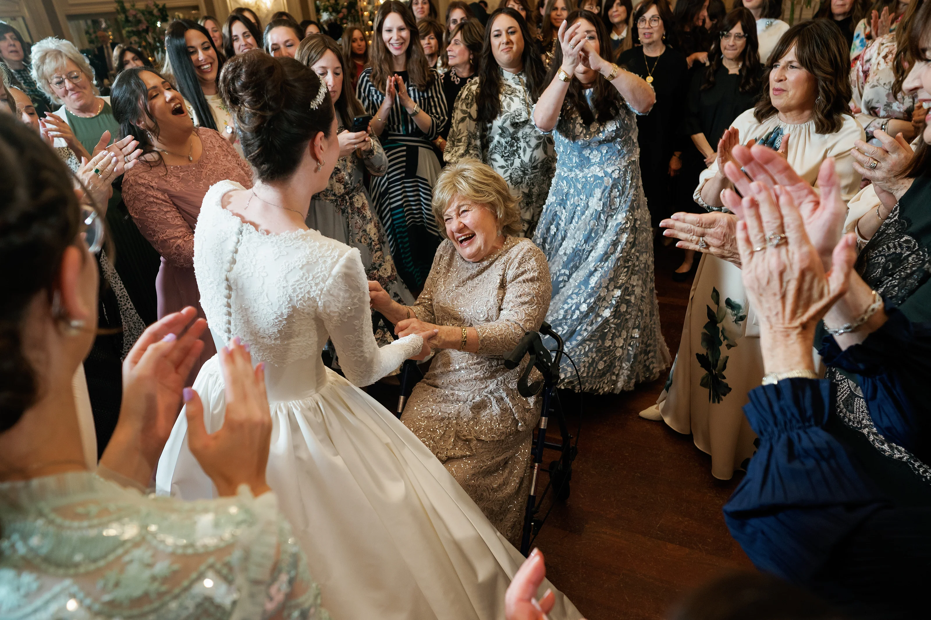 Bride in a white gown dancing as guests in the background clap and cheer, with a joyful close-up of an older woman smiling beside the aisle.
