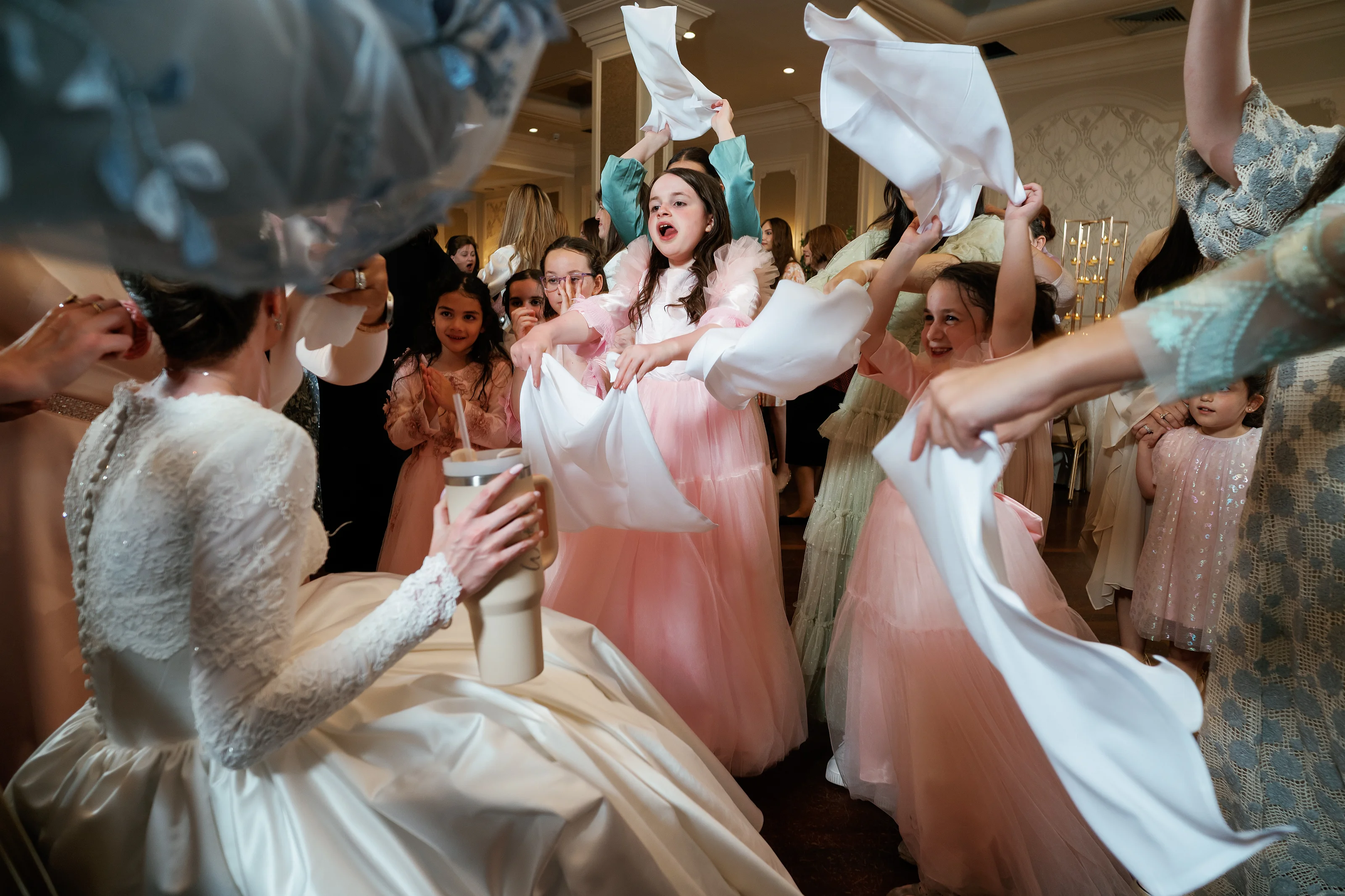 A group of little girls in pastel pink and white dresses dance in a ballroom, arms raised and smiling during a celebration.