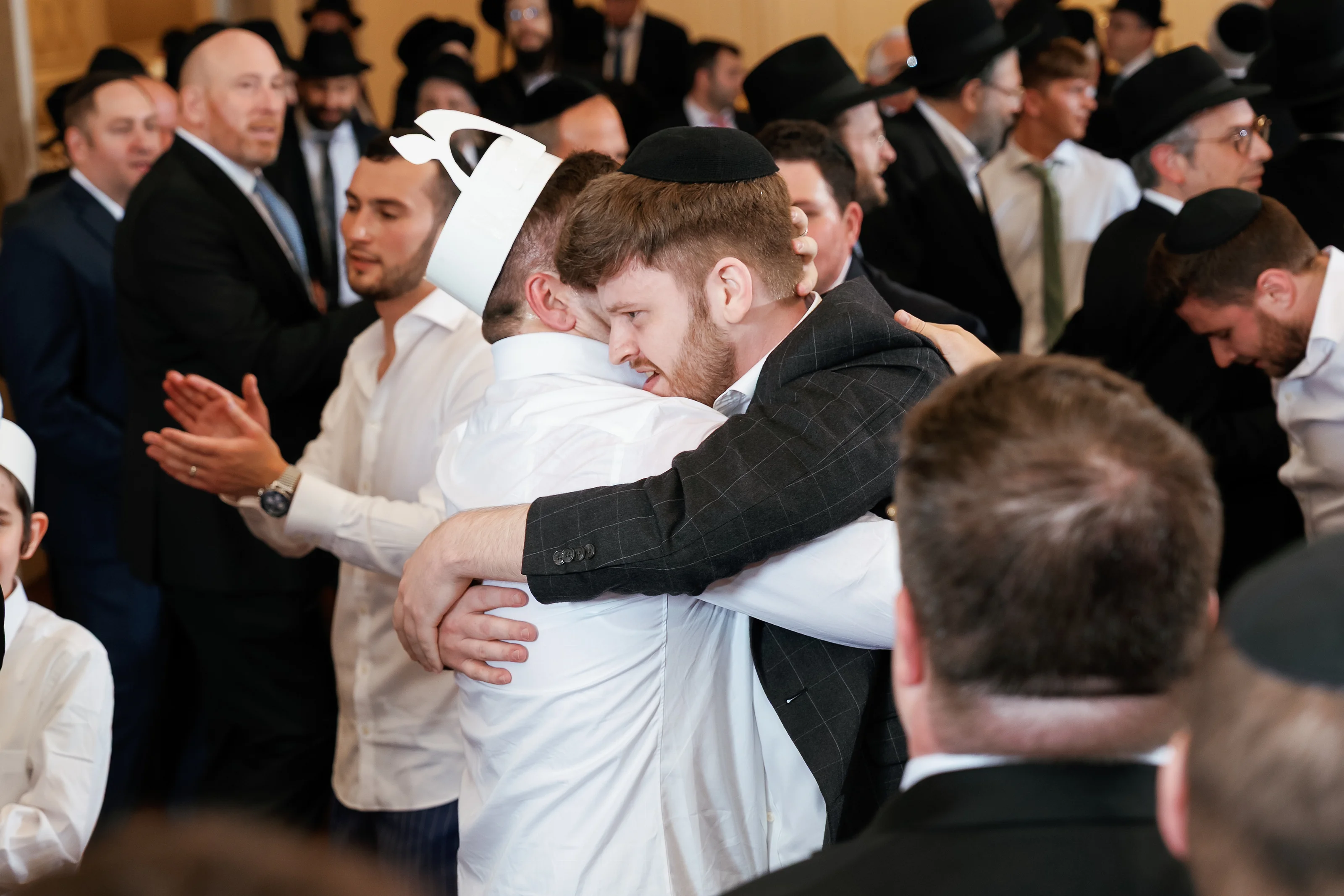 Candid embrace during a Jewish wedding celebration with the groom hugging another man while guests in formal hats clap in the background.