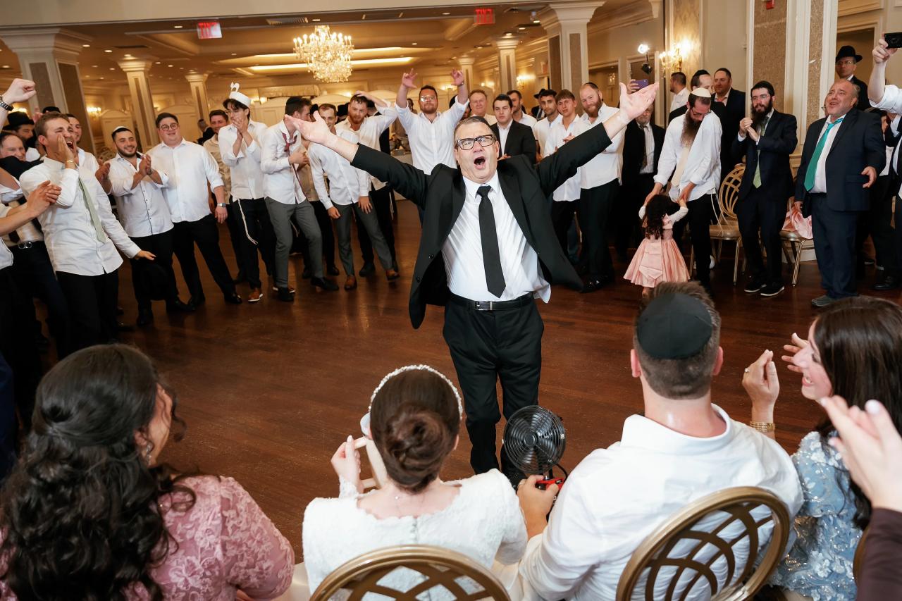A celebrant in a suit leads an energetic dance on the wedding reception floor with guests cheering in the background and arms raised.
