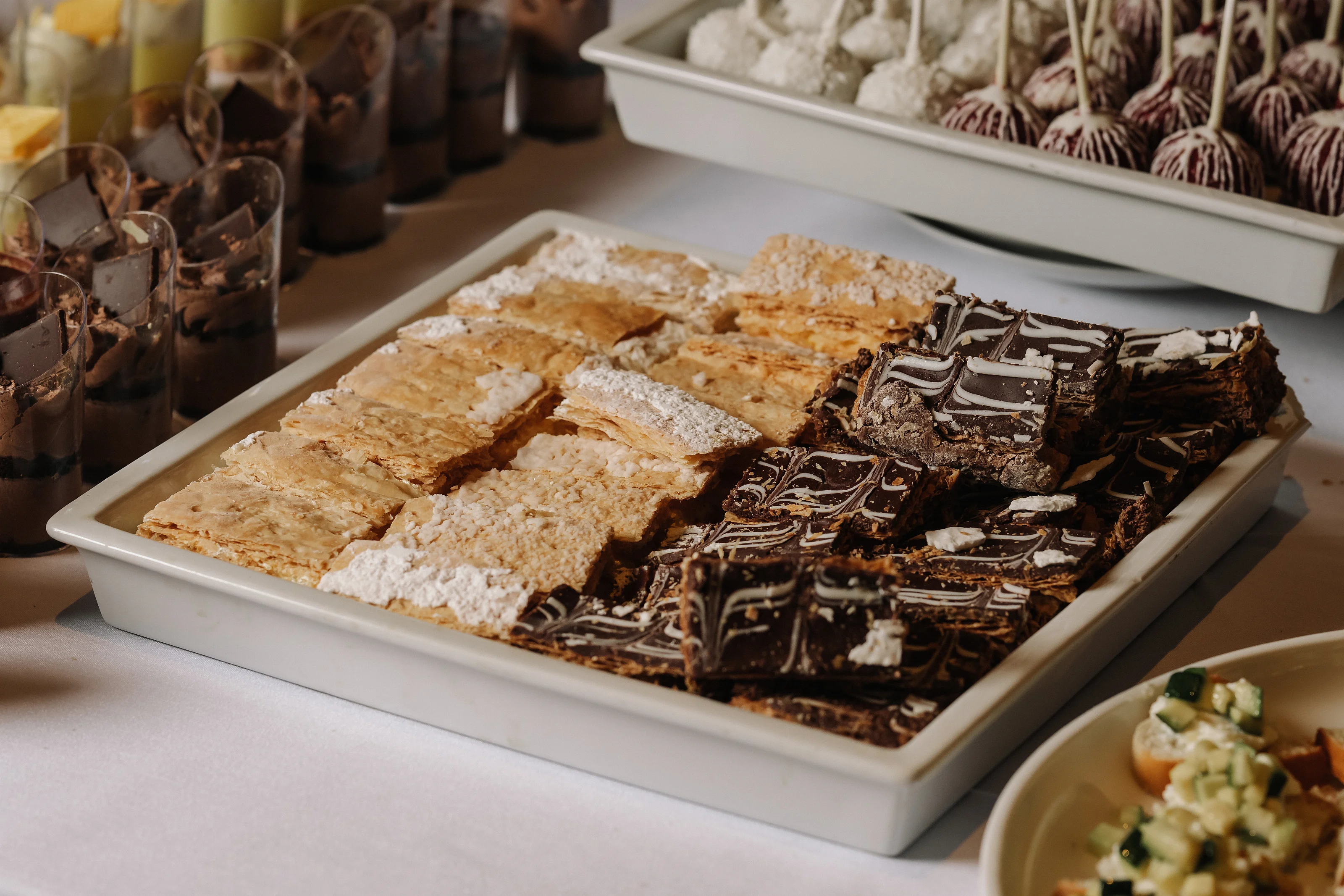 Close-up of an assortment of desserts in a white serving tray with chocolate-topped pieces and warm reception lighting