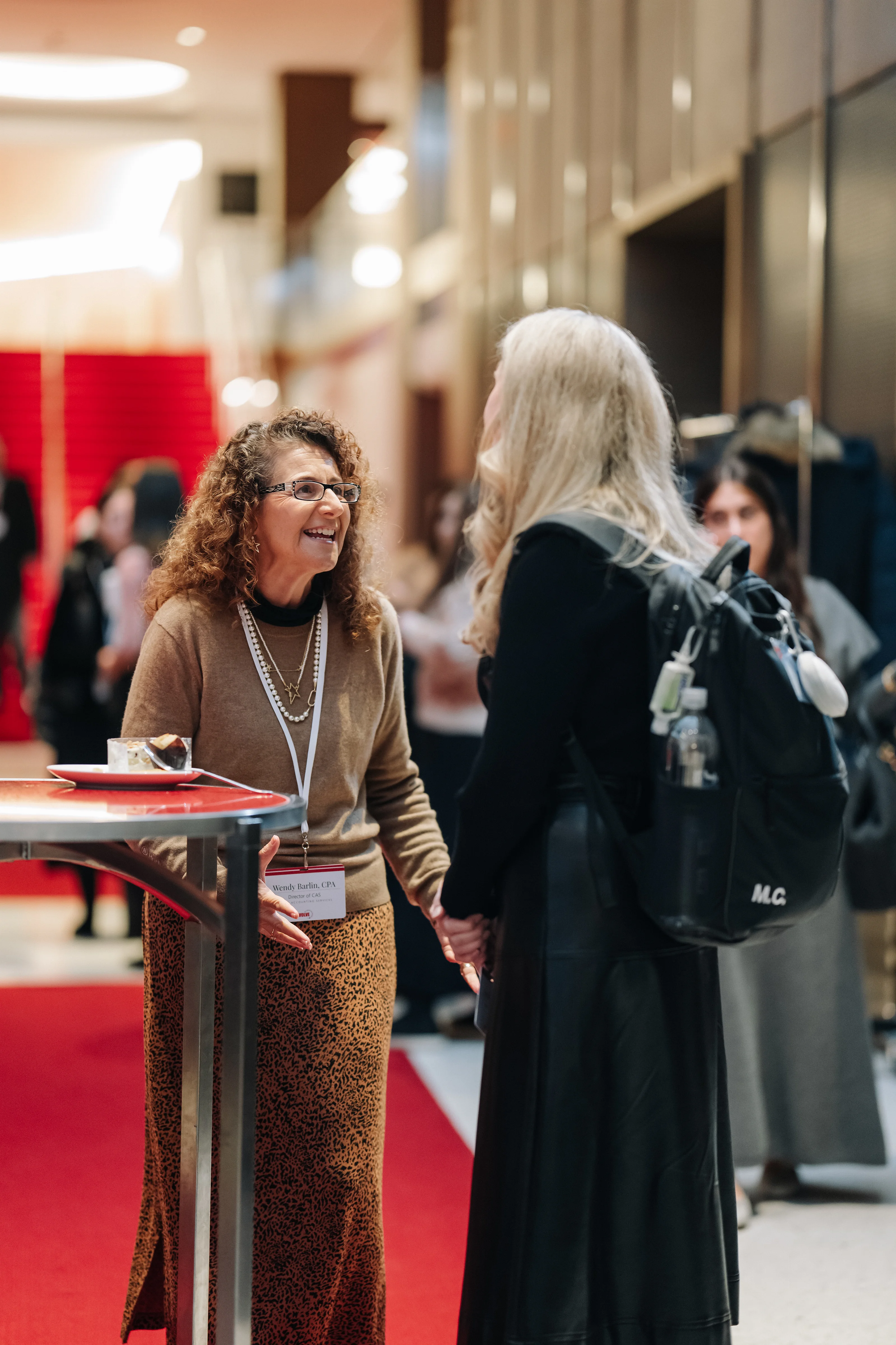 Smiling woman conversing near a red reception table on an indoor wedding event with a red carpet and blurred guests behind her.