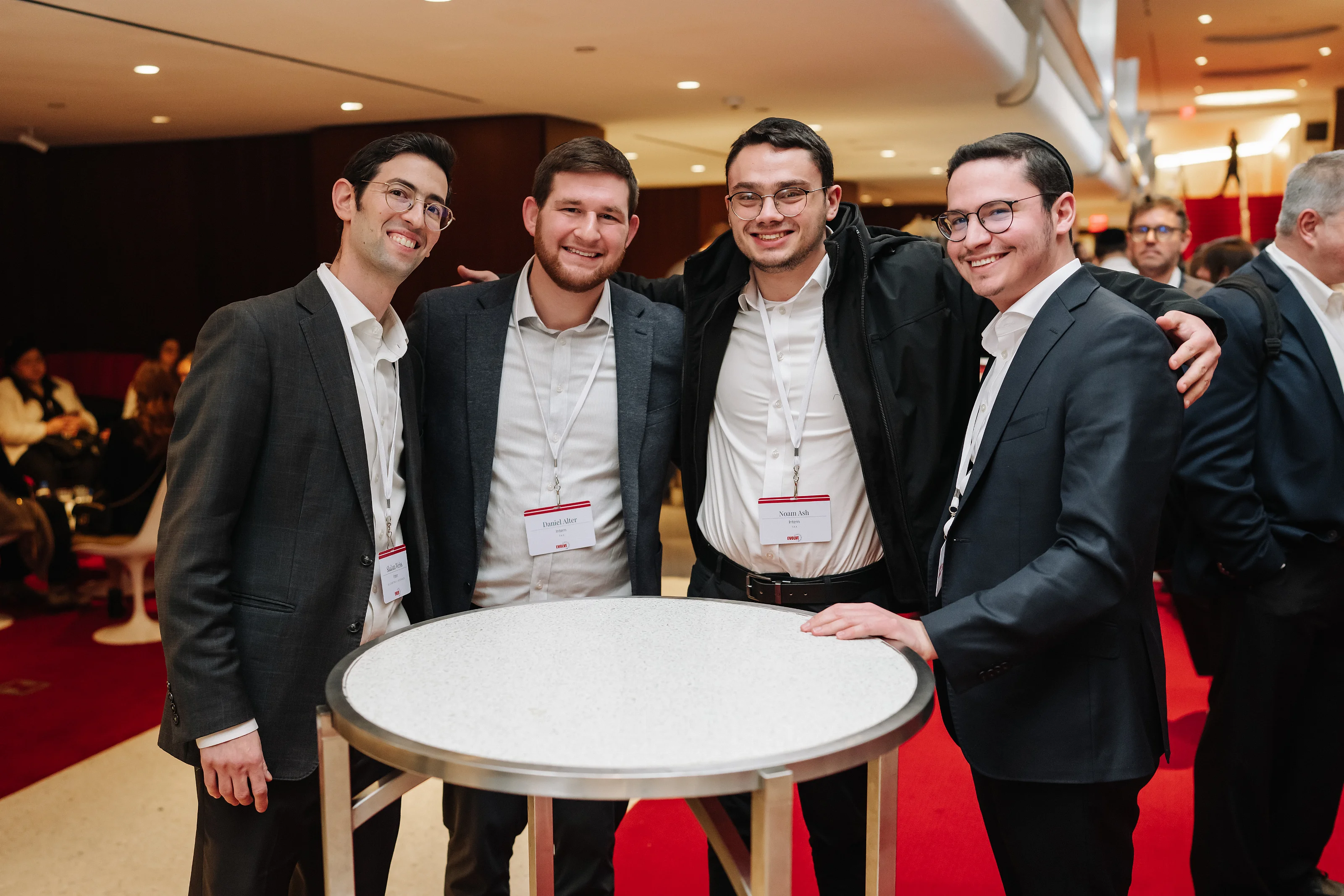 Four men in suits smiling together indoors around a small round table on a red-carpet event floor.
