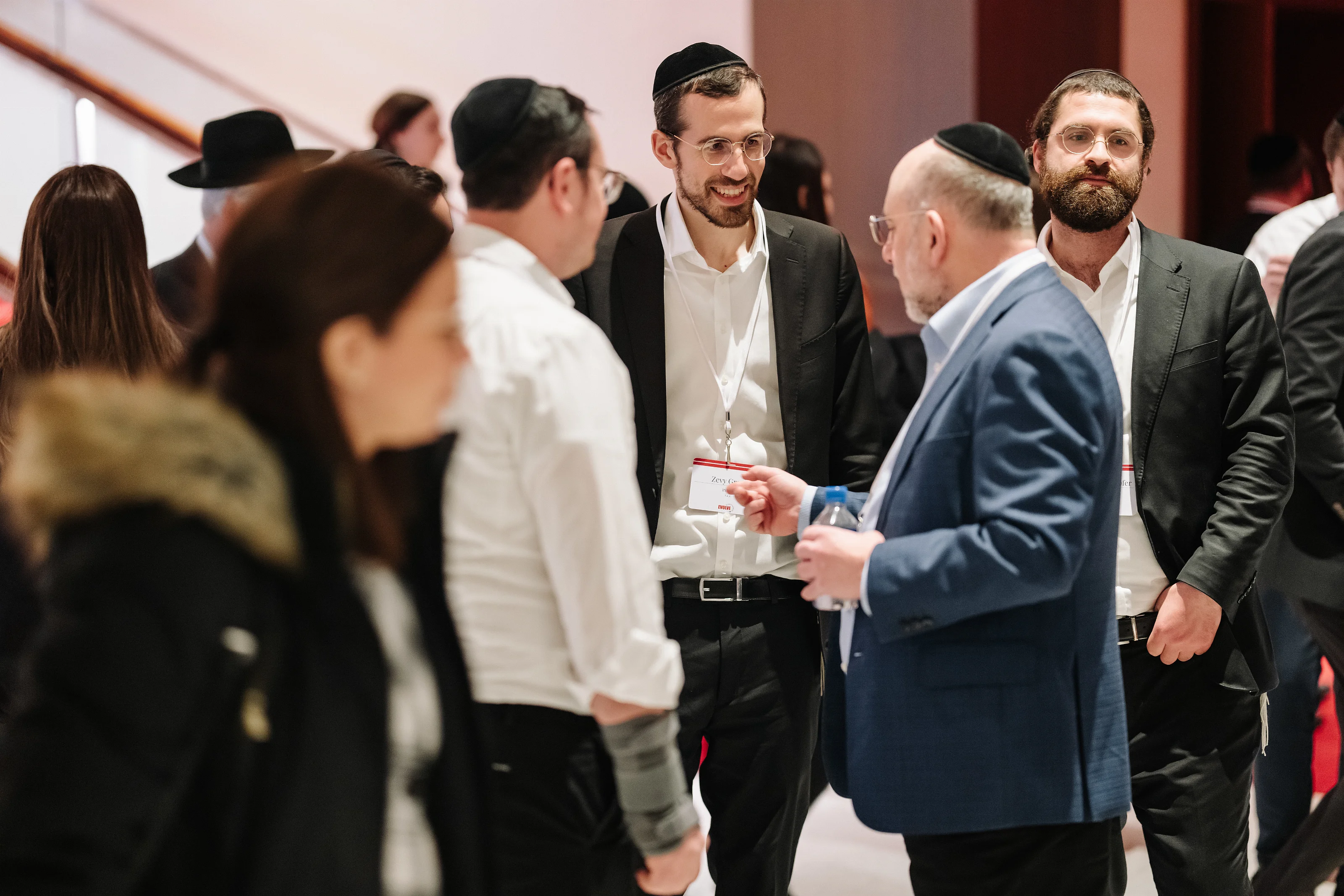 Smiling man in formal attire and kippah in focus while other guests are blurred at an indoor wedding event