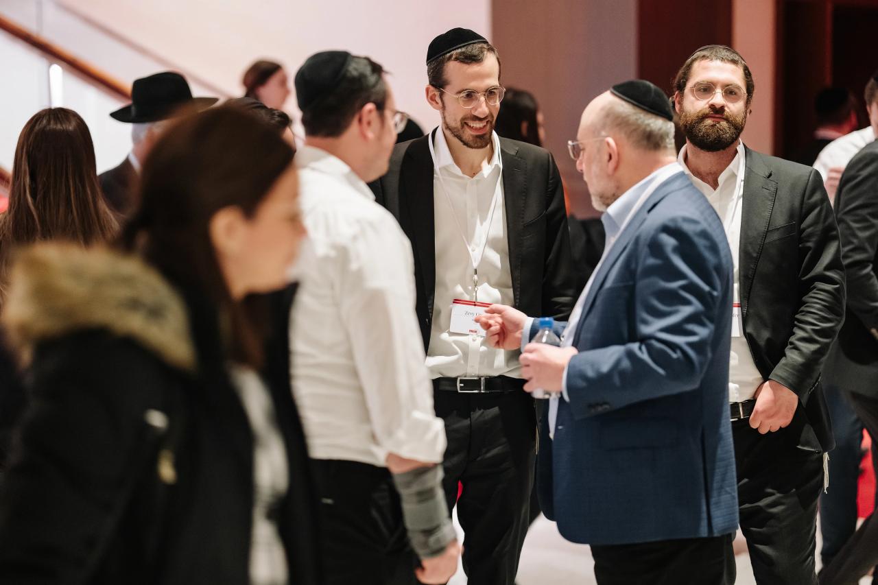 Smiling man in formal attire and kippah in focus while other guests are blurred at an indoor wedding event