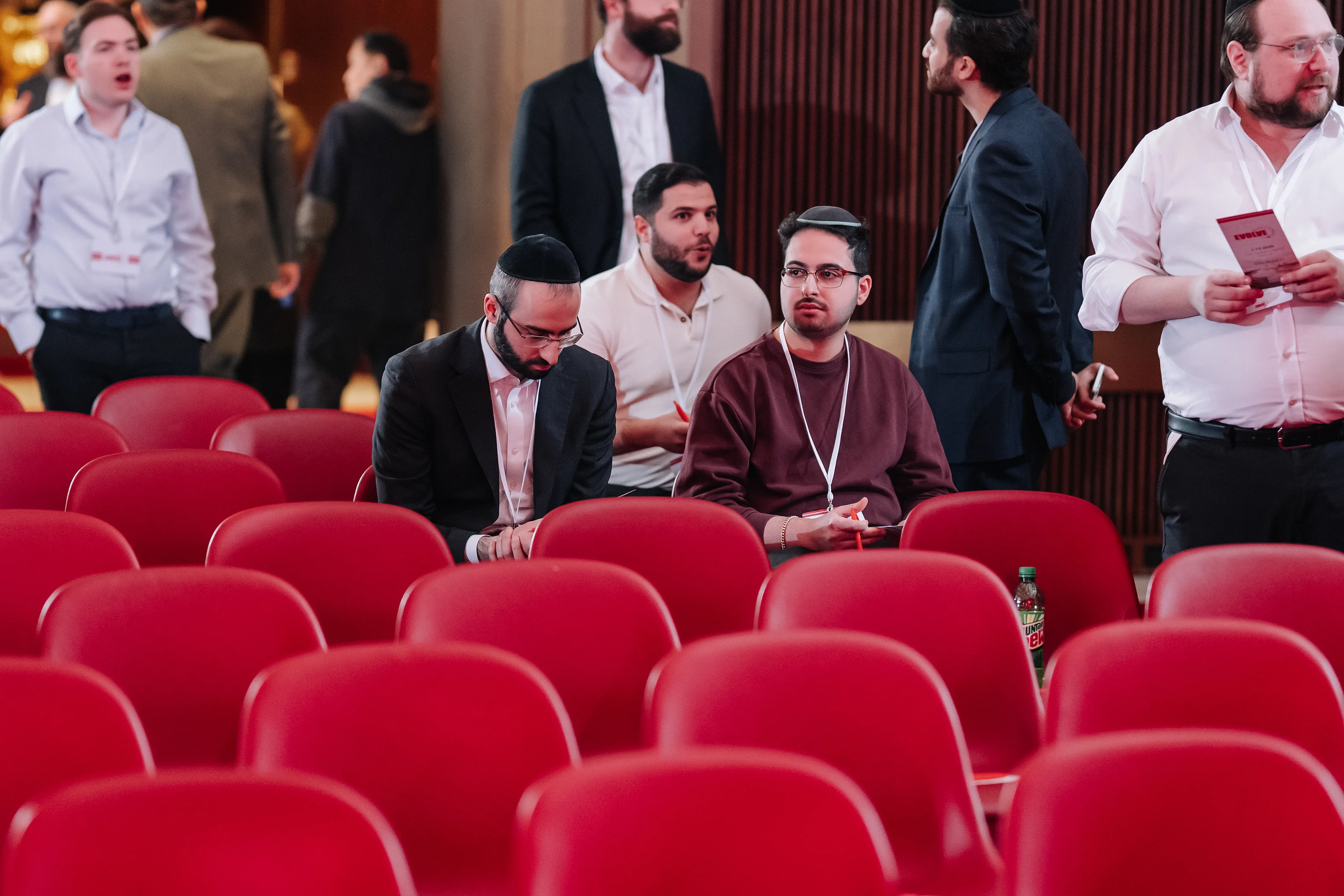 Two seated men with yarmulks and lanyards in rows of red theater seats during an indoor Jewish event, with attendees in the background.